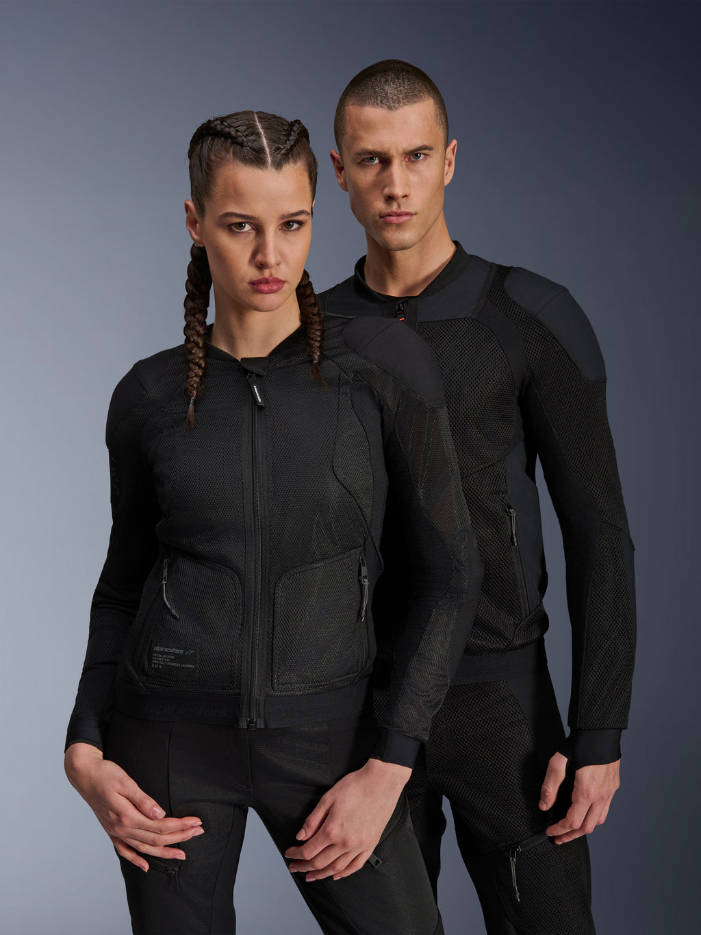 Hombre y mujer con chaquetas Alpinestars Troop-Air y Stella Troop-Air, chaquetas de moto de malla técnica negras, con estructura de malla técnica 2D para ventilación y paneles reforzados, ropa de conducción urbana contemporánea.