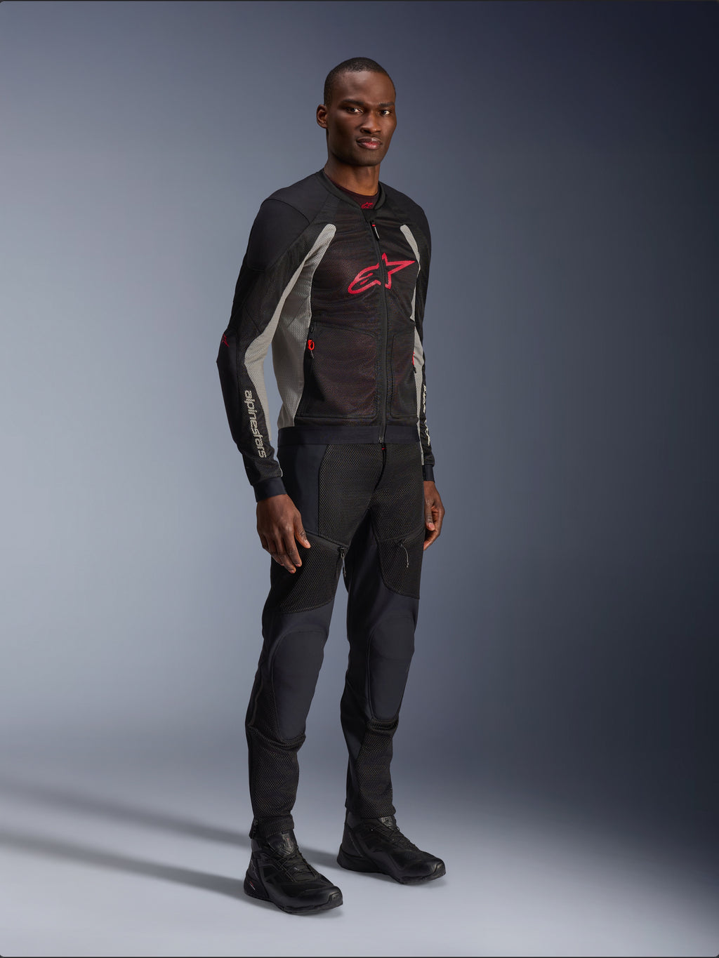 Chaqueta Alpinestars Troop-Air, chaqueta de malla técnica para motocicleta en negro, gris ceniza y rojo brillante, usada por un modelo con pantalones y botas negros a juego, con un logotipo de Alpinestars rojo en el pecho, equipamiento de conducción urbano contemporáneo.