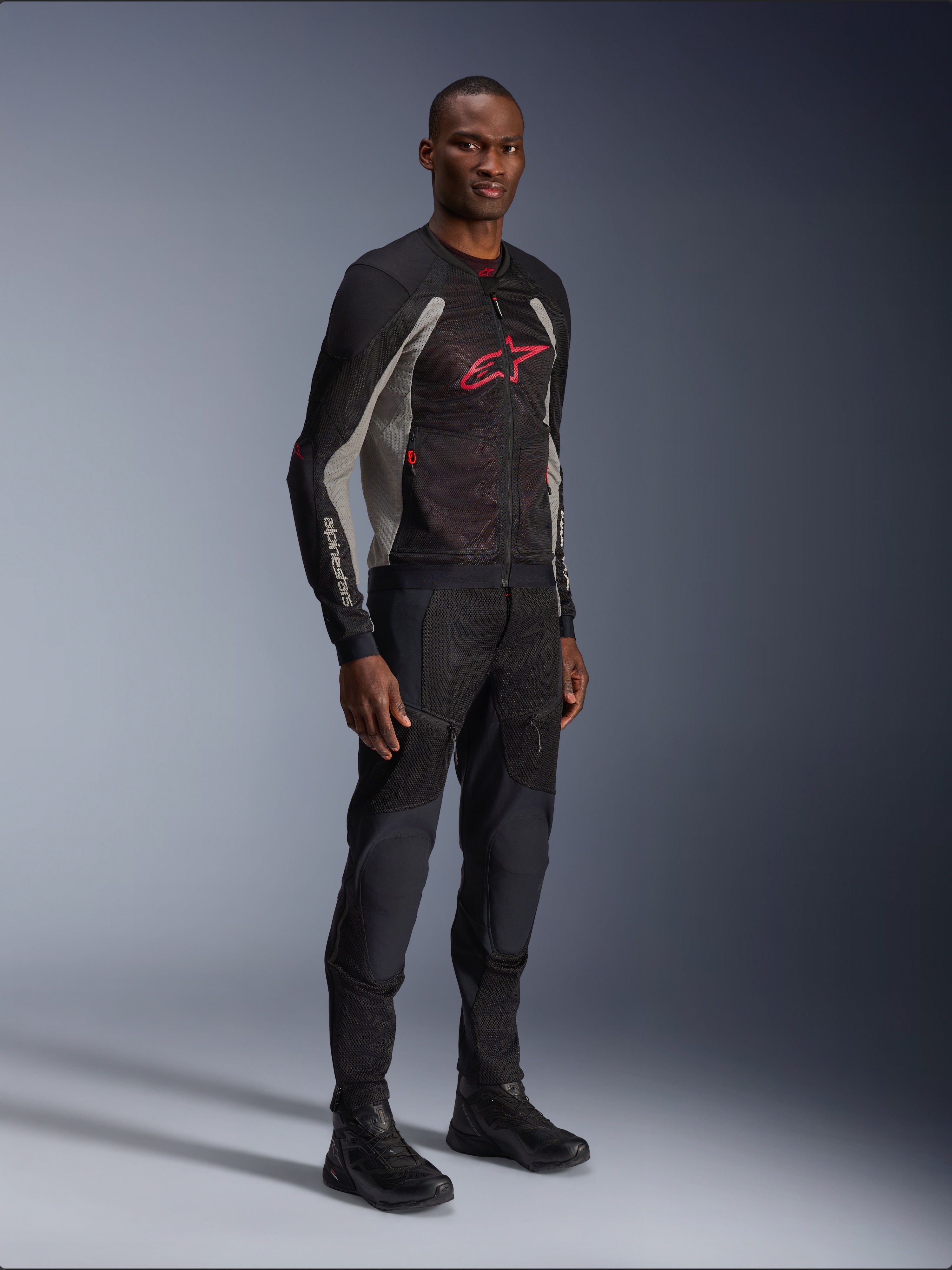 Chaqueta Alpinestars Troop-Air, chaqueta de malla técnica para motocicleta en negro, gris ceniza y rojo brillante, usada por un modelo con pantalones y botas negros a juego, con un logotipo de Alpinestars rojo en el pecho, equipamiento de conducción urbano contemporáneo.