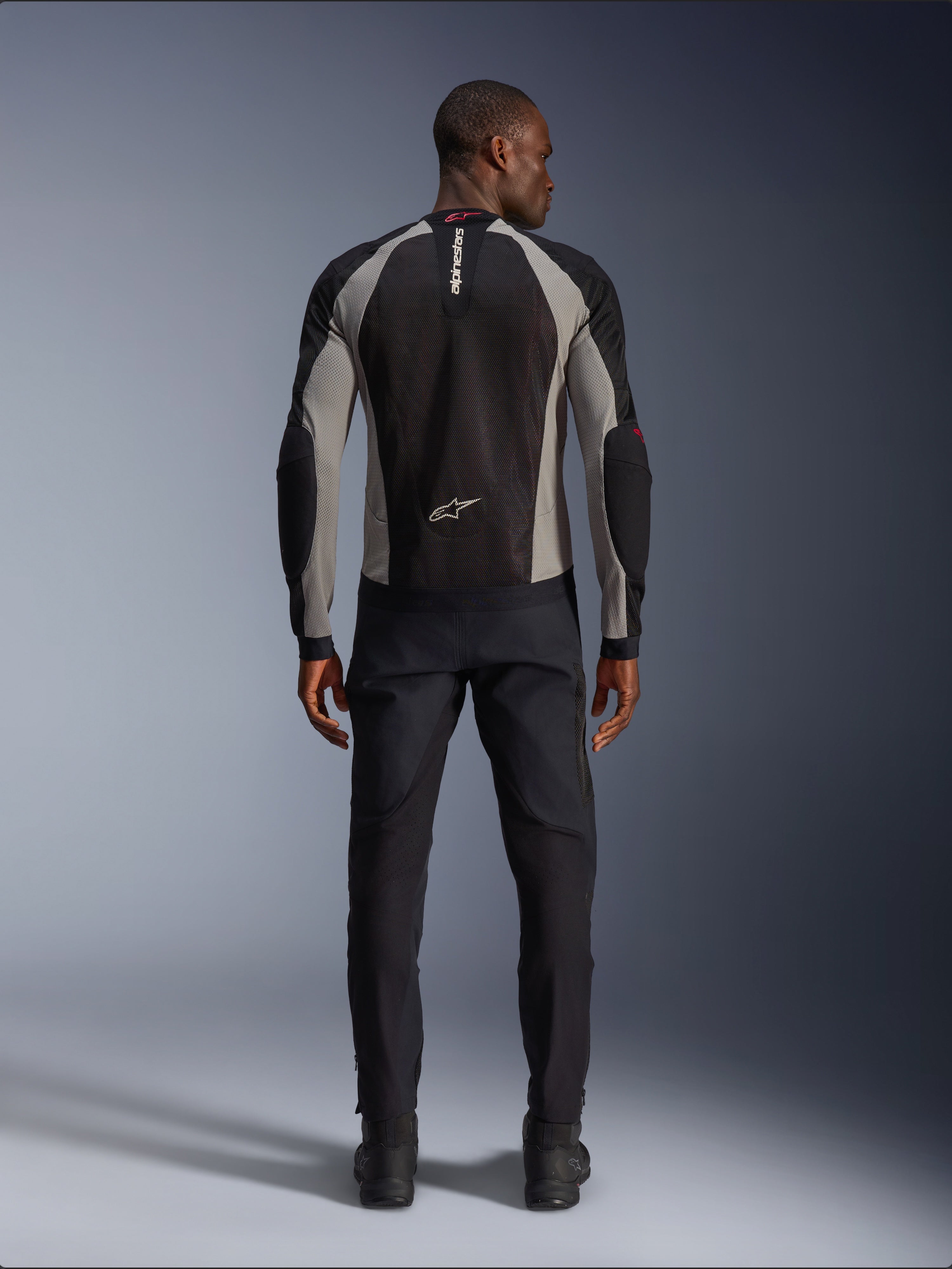 Chaqueta Alpinestars Troop-Air, chaqueta de moto negra, gris ceniza y rojo brillante, vista trasera en modelo masculino, construcción de malla ligera para ventilación, marca Alpinestars en el cuello y la parte inferior de la espalda, combinada con pantalones de conducción negros