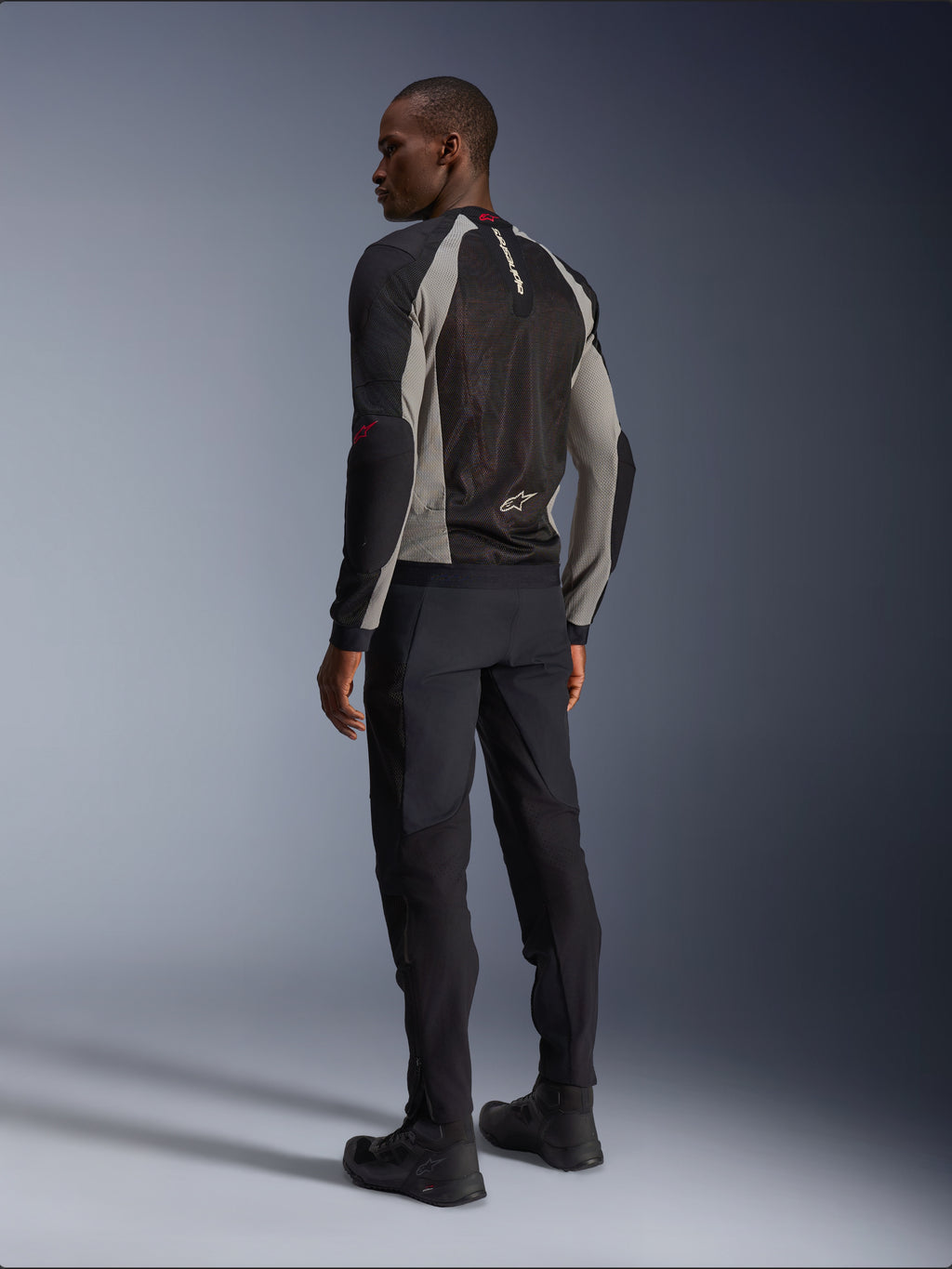 Modelo luciendo la chaqueta Alpinestars Troop-Air vista desde atrás, en color negro y gris ceniza con detalles en rojo brillante, construcción de malla técnica para ventilación, combinada con pantalones de moto negros y botas de conducción, estilo urbano contemporáneo