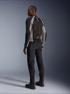 Modelo luciendo la chaqueta Alpinestars Troop-Air vista desde atrás, en color negro y gris ceniza con detalles en rojo brillante, construcción de malla técnica para ventilación, combinada con pantalones de moto negros y botas de conducción, estilo urbano contemporáneo