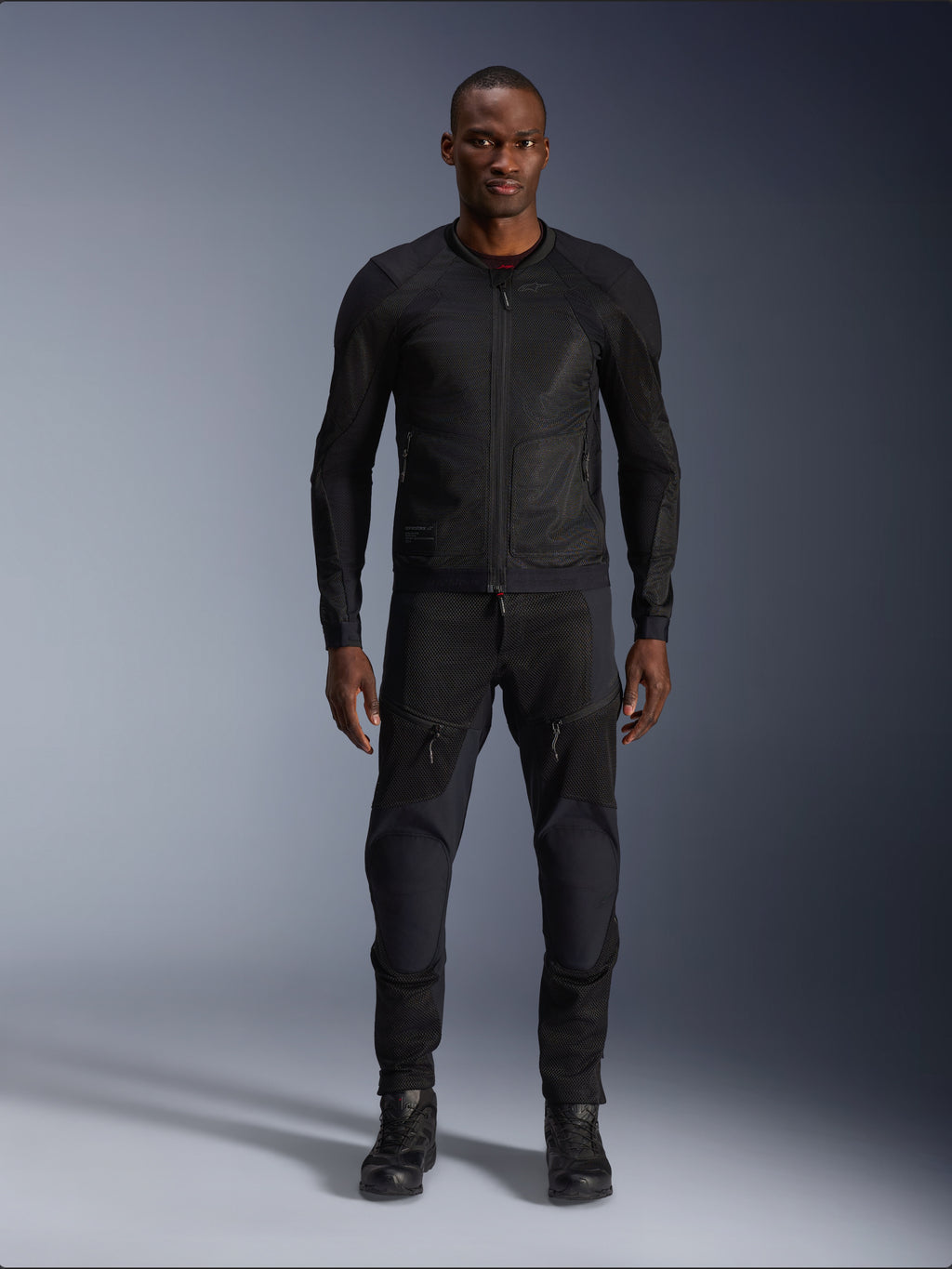 Modelo masculino vistiendo la chaqueta Alpinestars Troop-Air en color Black Black, chaqueta de moto de malla ligera, combinada con pantalones y botas de montar negros a juego, con una construcción técnica de malla 2D para un flujo de aire superior.