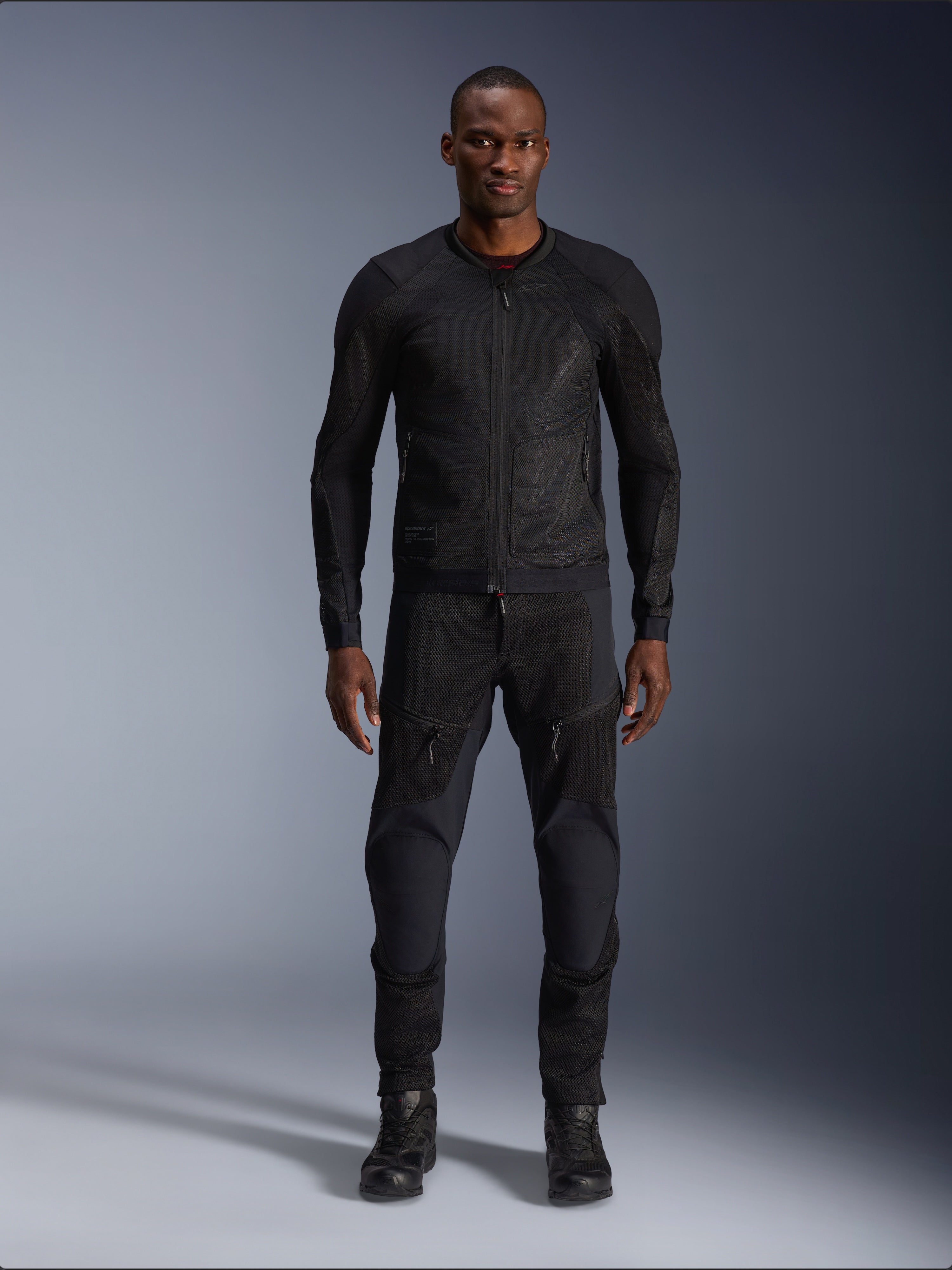 Modelo masculino vistiendo la chaqueta Alpinestars Troop-Air en color Black Black, chaqueta de moto de malla ligera, combinada con pantalones y botas de montar negros a juego, con una construcción técnica de malla 2D para un flujo de aire superior.