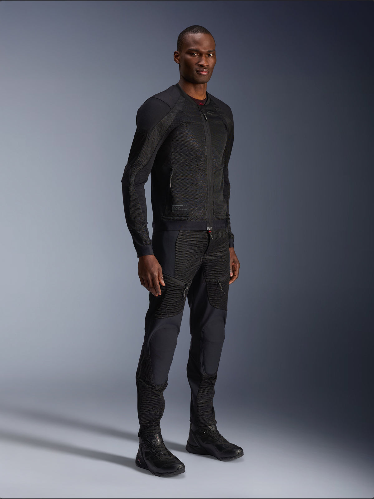Alpinestars Troop-Air Jacket, negra, chaqueta técnica de malla para motociclismo usada por un modelo con pantalones y botas negros a juego, con construcción de malla ligera y diseño urbano minimalista