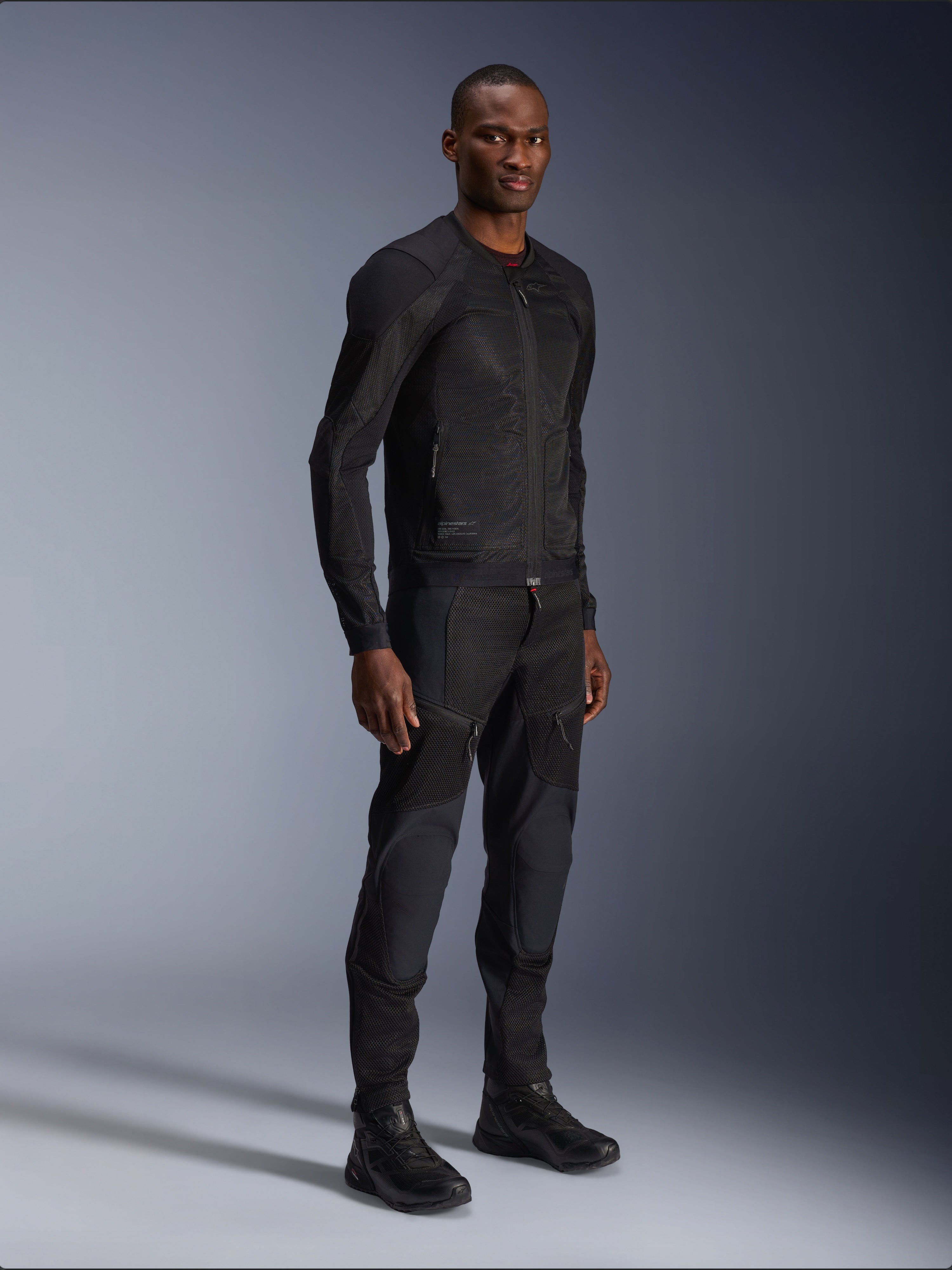 Alpinestars Troop-Air Jacket, negra, chaqueta técnica de malla para motociclismo usada por un modelo con pantalones y botas negros a juego, con construcción de malla ligera y diseño urbano minimalista