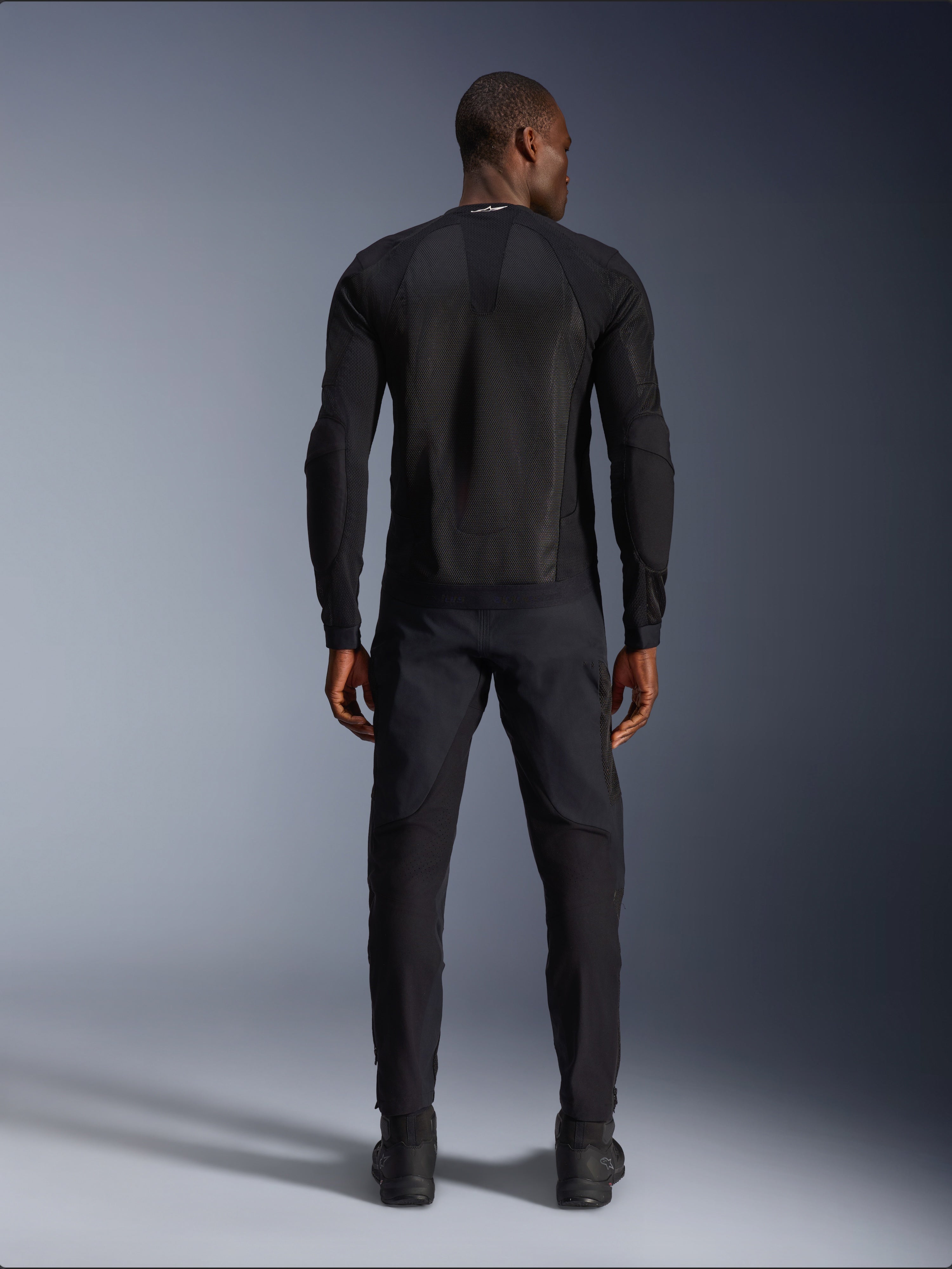 Chaqueta Alpinestars Troop-Air en Negro Negro, vista trasera que muestra el panel de malla técnica en la espalda y su construcción ventilada, combinada con pantalones de motociclista y botas negras a juego