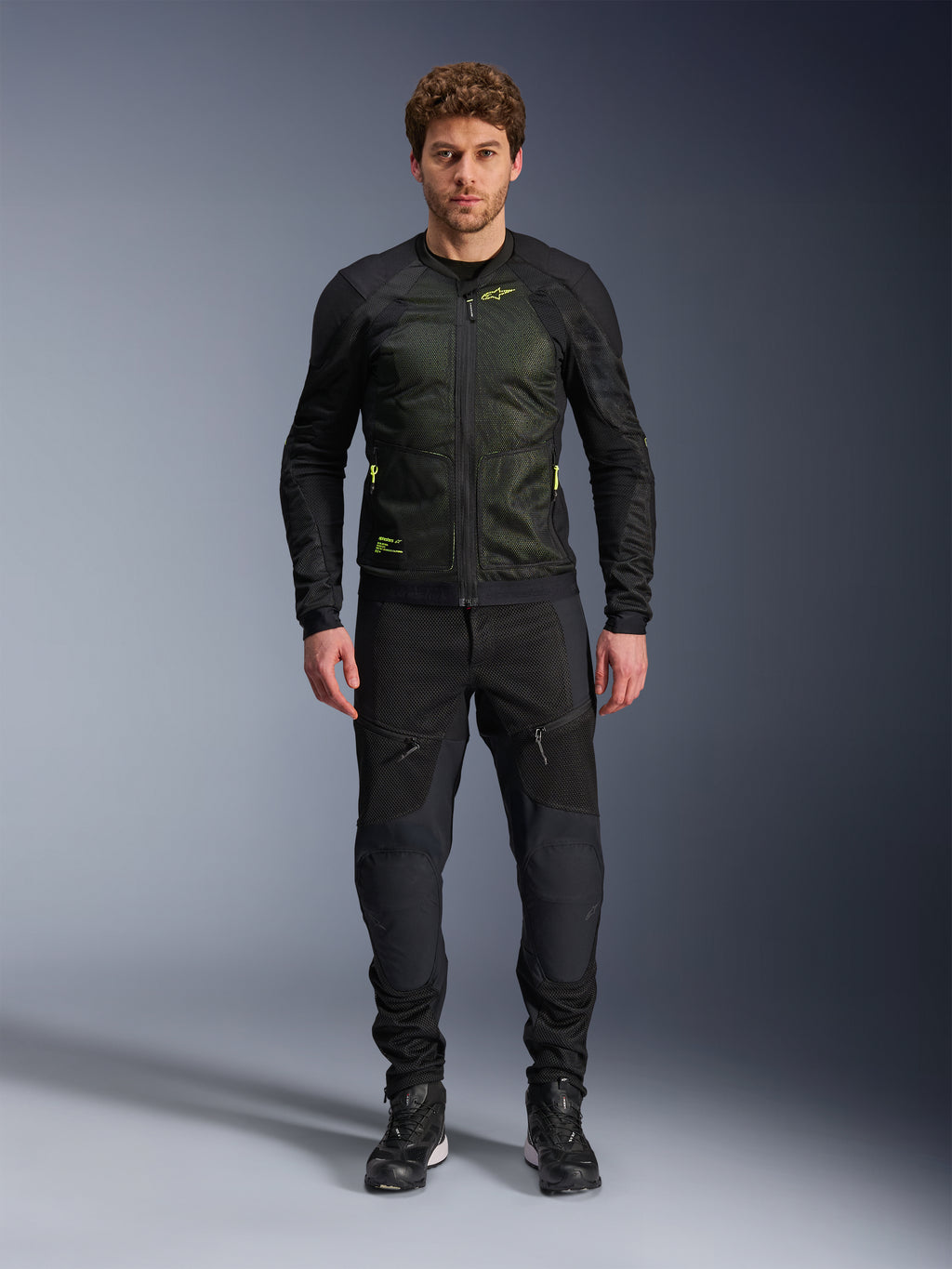 Modelo masculino vistiendo la chaqueta Alpinestars Troop-Air en Negro Amarillo Flúor, con paneles de malla técnica y detalles en amarillo flúor, combinada con pantalones y calzado de motociclismo negros, equipamiento contemporáneo para climas cálidos