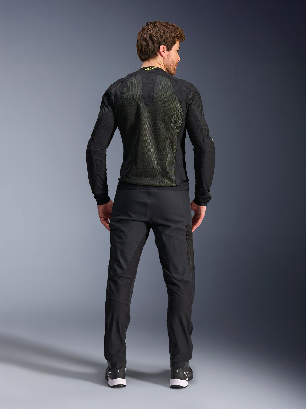 Chaqueta Alpinestars Troop-Air, Negro Amarillo Flúor, vista trasera en un modelo, con un gran panel posterior de malla técnica 2D para ventilación, chaqueta técnica de motociclismo usada con pantalones y botas de conducción negros, estilo de moto urbano