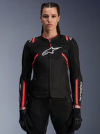 Chaqueta para mujer Alpinestars Stella T-Sps Air V2, negro y rosa diva, chaqueta textil para moto con paneles de malla transpirable y detalles en rosa, logo blanco de Alpinestars en el pecho, usada por una modelo con pantalones y guantes de motociclismo negros