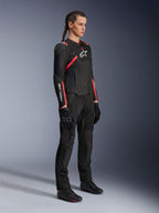 Piloto femenina con la chaqueta Alpinestars Stella T-SPS Air V2 en negro y rosa diva, chaqueta de moto textil para mujer con paneles de malla y acentos en rosa, combinada con pantalones de conducción y guantes negros, vista de cuerpo completo en estudio.