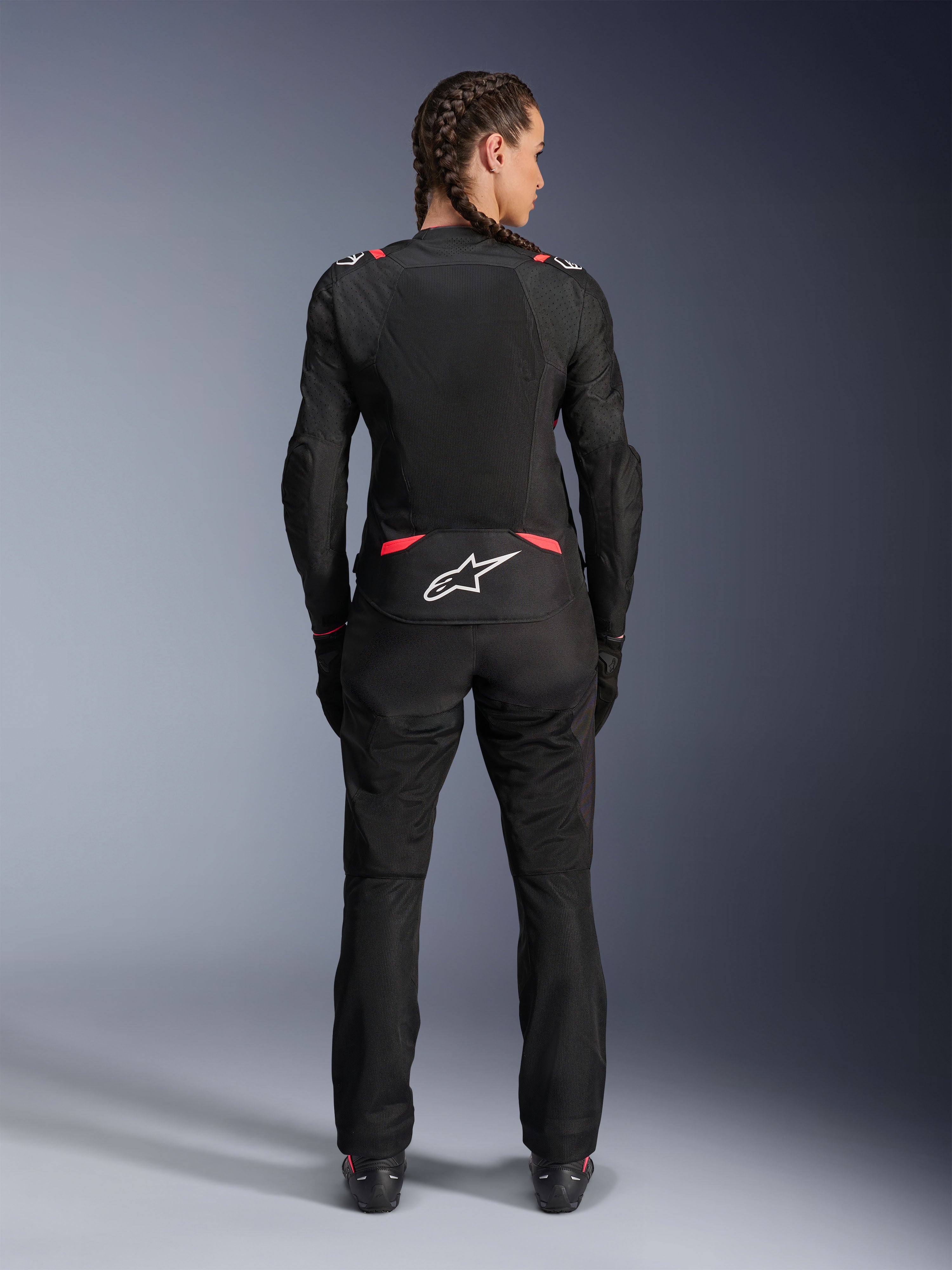 Chaqueta Alpinestars Stella T-SPS Air V2, chaqueta de moto textil para mujer, negro y rosa Diva, vista posterior en modelo con cabello trenzado, con grandes paneles de malla para ventilación, logo Astars blanco en la parte inferior de la espalda, combinada con pantalones de moto negros a juego.