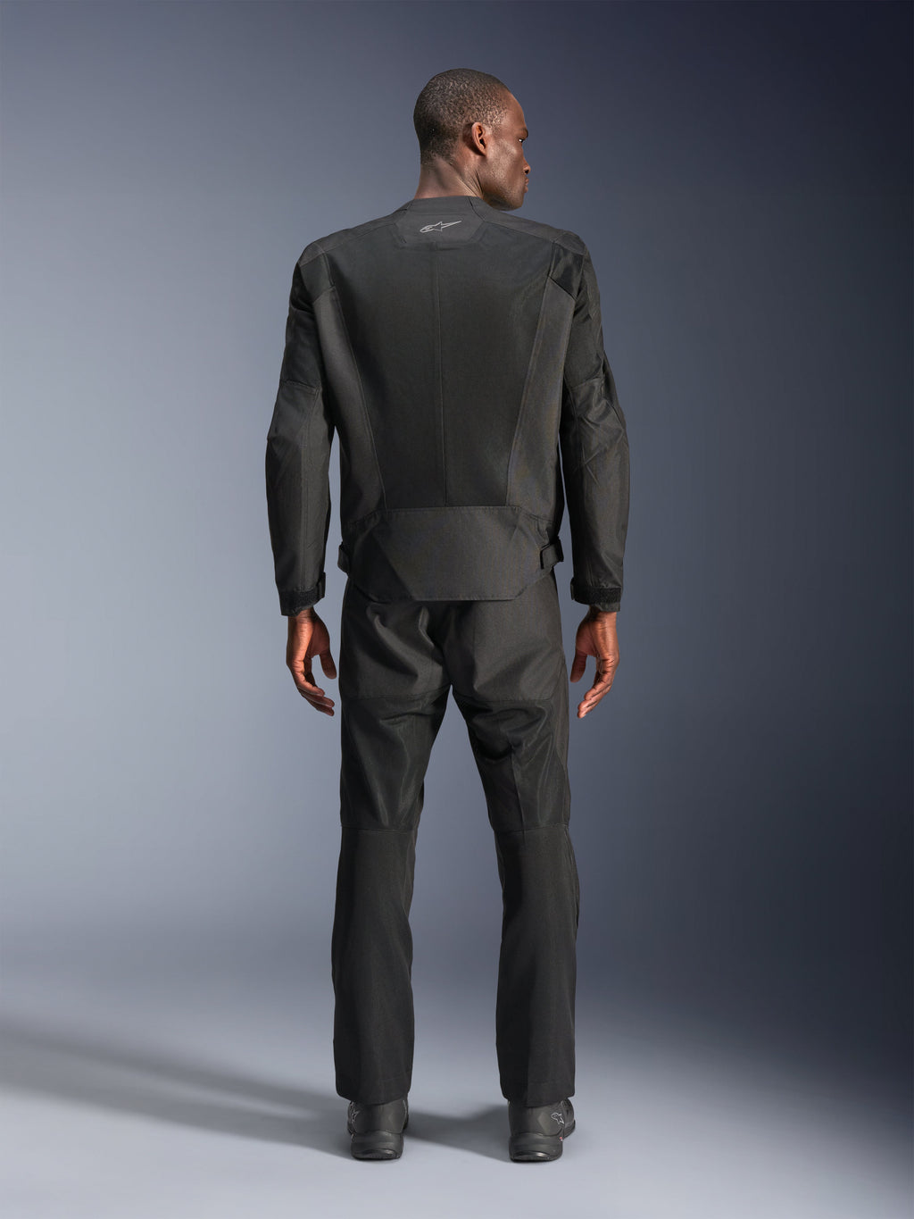 Vista posterior de un hombre con la chaqueta Alpinestars C-1 Air en color negro, chaqueta de moto urbana con paneles de malla técnica, pantalones textiles y botas negras a juego, estética limpia con logotipo reflectante en la parte superior de la espalda.