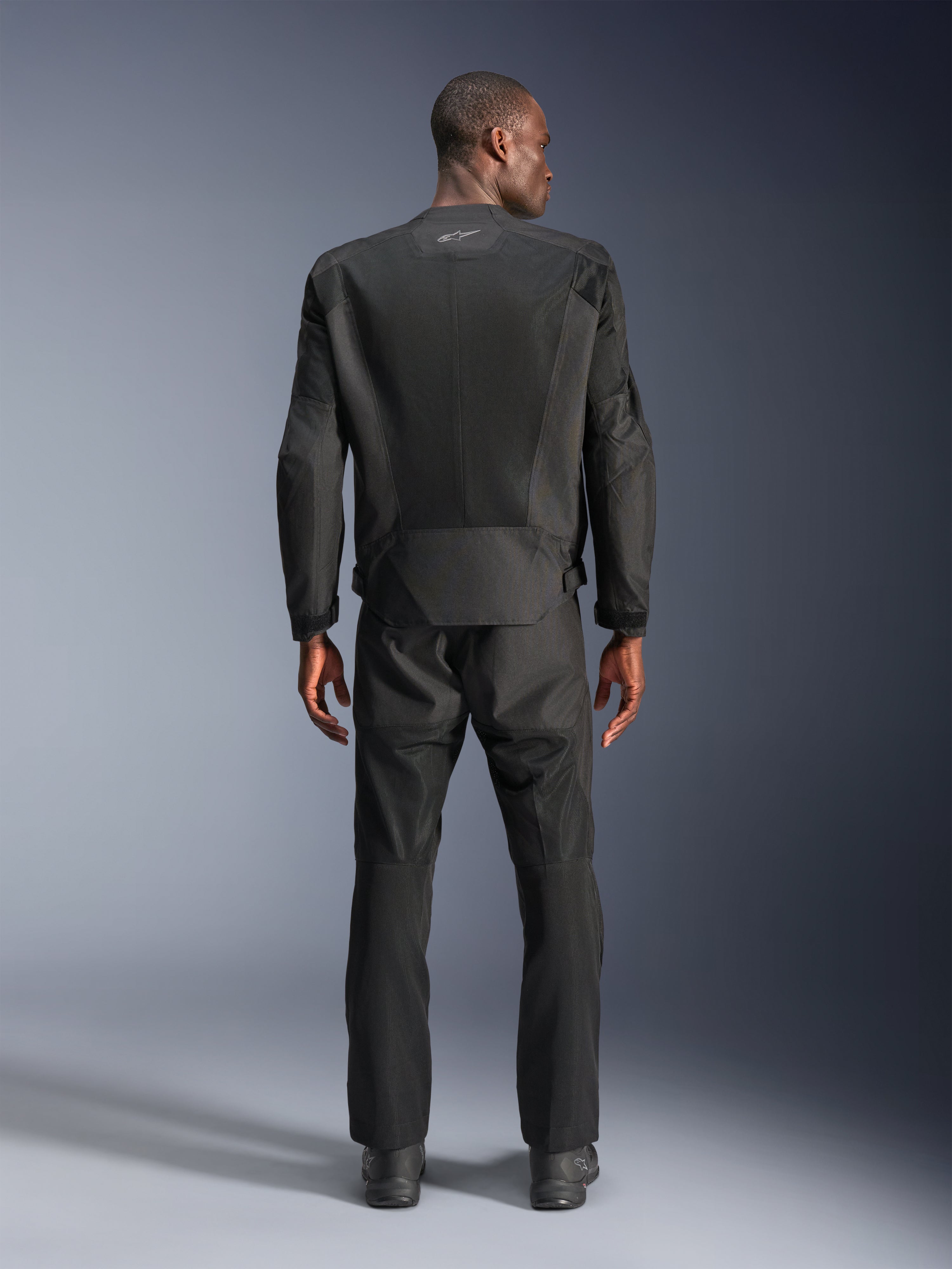 Vista posterior de un hombre con la chaqueta Alpinestars C-1 Air en color negro, chaqueta de moto urbana con paneles de malla técnica, pantalones textiles y botas negras a juego, estética limpia con logotipo reflectante en la parte superior de la espalda.