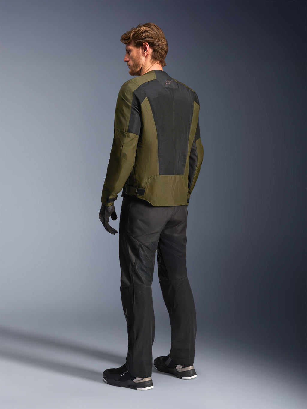 Modelo masculino con la chaqueta Alpinestars C-1 Air en color kaki, chaqueta de moto urbana, vista trasera destacando los grandes paneles de malla negra en la espalda, combinada con pantalones técnicos de conducción y guantes negros, equipamiento de moto contemporáneo para clima cálido