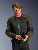 Hombre vistiendo la chaqueta Alpinestars C-1 Air en Khaki, chaqueta de moto urbana con tejido verde oliva y paneles de malla negra, mirando hacia abajo mientras sostiene guantes de moto negros, estilo limpio y contemporáneo para conducción en carretera.
