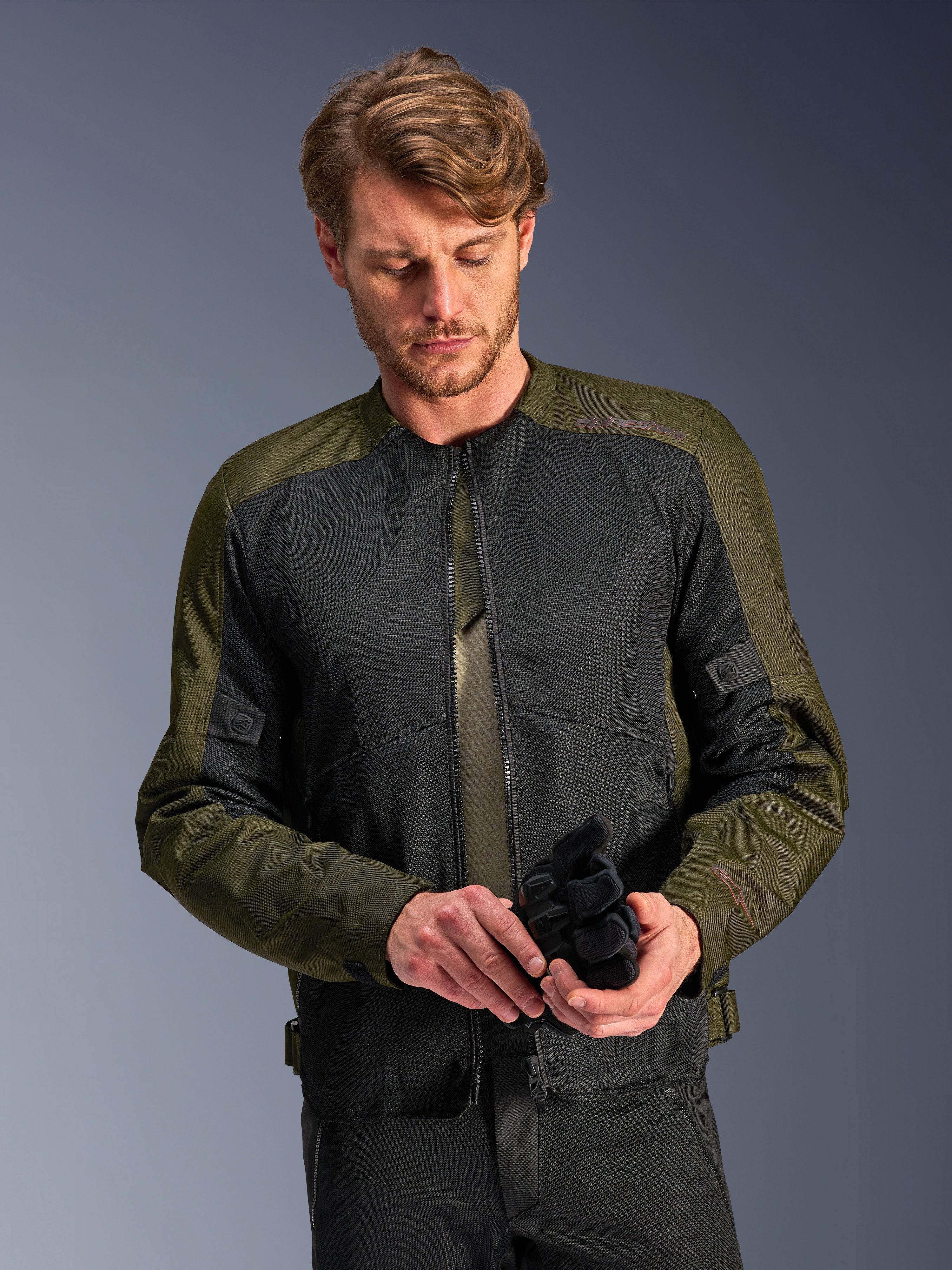 Hombre vistiendo la chaqueta Alpinestars C-1 Air en Khaki, chaqueta de moto urbana con tejido verde oliva y paneles de malla negra, mirando hacia abajo mientras sostiene guantes de moto negros, estilo limpio y contemporáneo para conducción en carretera.