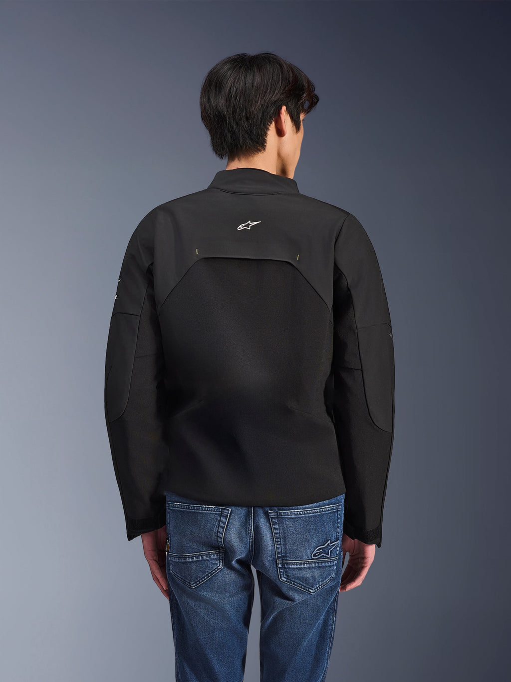 Chaqueta Alpinestars Provoke, chaqueta de moto urbana, negra, vista trasera que muestra el logo blanco de Astars y paneles técnicos de ripstop, combinada con pantalones vaqueros azules, diseño contemporáneo para desplazamientos urbanos