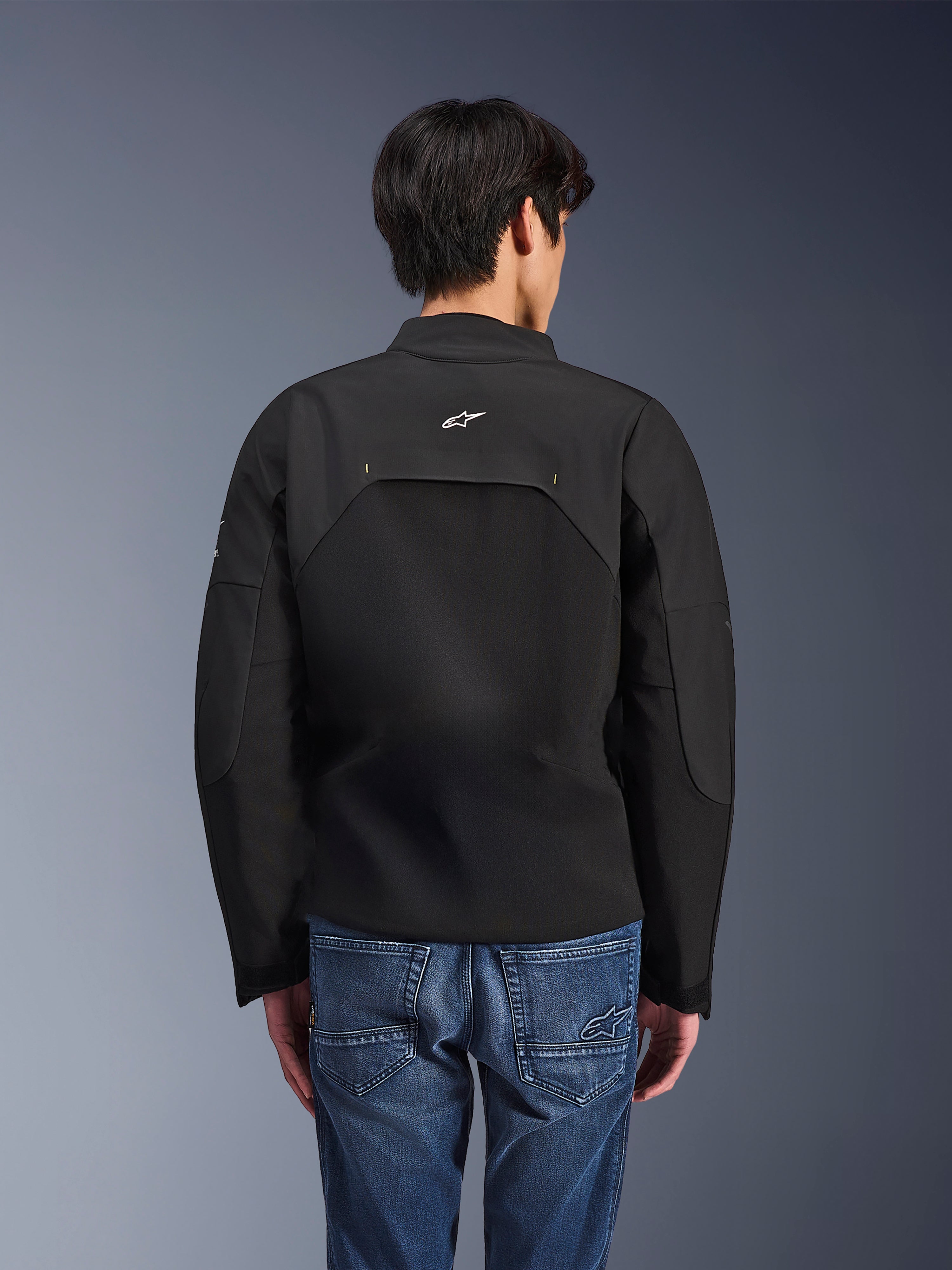 Chaqueta Alpinestars Provoke, chaqueta de moto urbana, negra, vista trasera que muestra el logo blanco de Astars y paneles técnicos de ripstop, combinada con pantalones vaqueros azules, diseño contemporáneo para desplazamientos urbanos