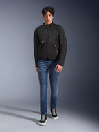 Modelo masculino vistiendo la chaqueta Alpinestars Provoke en color negro, chaqueta de moto textil urbana con cremalleras técnicas y paneles de softshell ripstop, combinada con vaqueros azules y zapatillas negras, estilo de motociclista urbano contemporáneo.