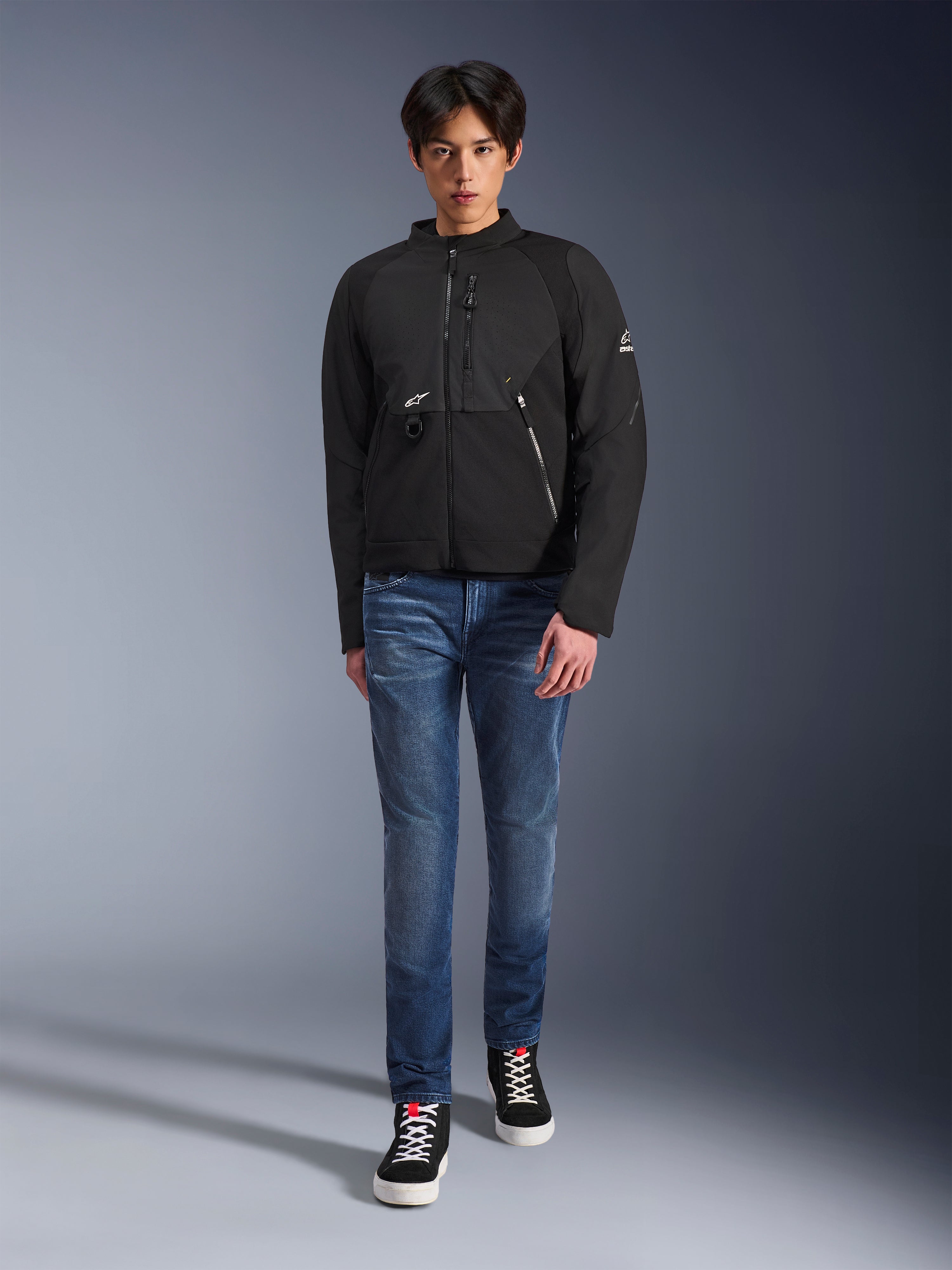 Modelo masculino vistiendo la chaqueta Alpinestars Provoke en color negro, chaqueta de moto textil urbana con cremalleras técnicas y paneles de softshell ripstop, combinada con vaqueros azules y zapatillas negras, estilo de motociclista urbano contemporáneo.