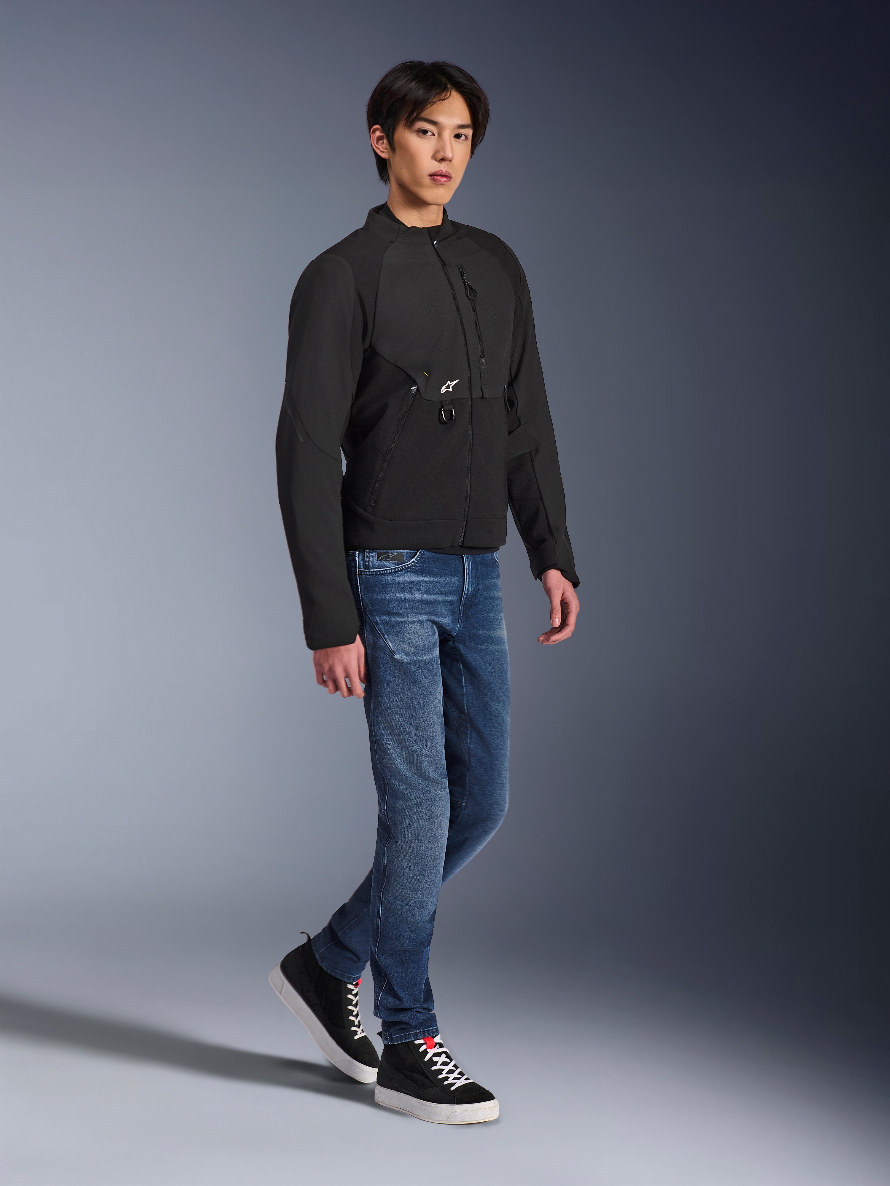 Chaqueta Alpinestars Provoke, chaqueta de moto urbana, negra, vestida por un modelo masculino con vaqueros azules y zapatillas negras, diseño textil técnico con un pequeño logotipo de Alpinestars, foto de estilo de vida de cuerpo completo para conducción urbana contemporánea.