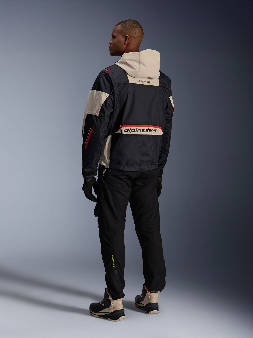 Vista trasera de un hombre con pantalones Alpinestars Acteon en color negro, combinados con una chaqueta Mogress Air en negro y crema, guantes negros y botas de conducción, conjunto de motociclismo urbano contemporáneo