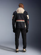 Rear view of a rider wearing the Alpinestars Mogress Air Jacket in Black, Almond, and Hot Orange, featuring a cream-colored hood, large Alpinestars branding across the back pocket, and orange zipper accents, paired with black pants and cream boots for an urban motorcycle look.
