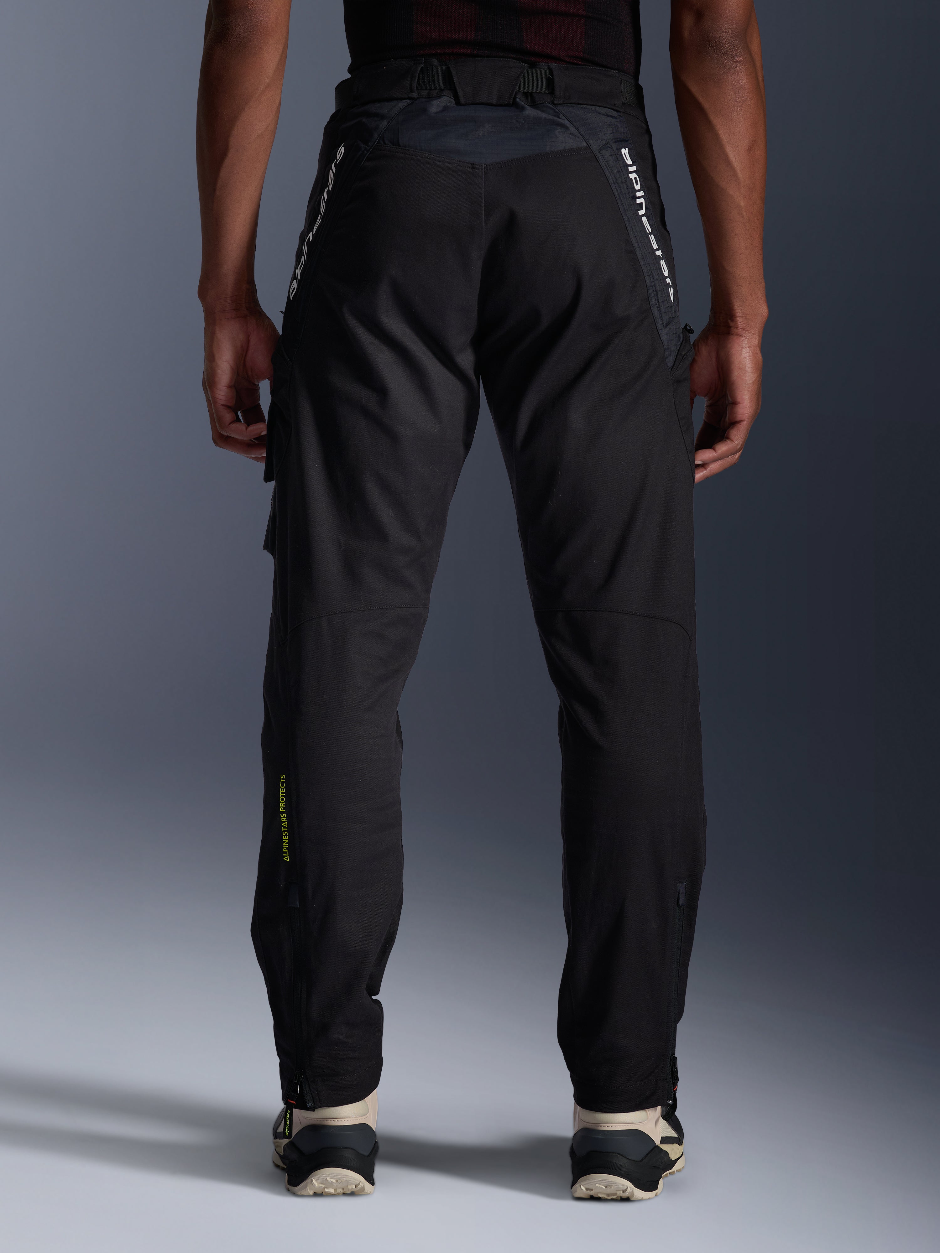Pantalones Alpinestars Acteon, pantalones urbanos de motociclismo, negros, vista trasera que muestra el ajuste entallado, logotipos blancos en la parte superior de las piernas y logotipo amarillo en la parte inferior de la pantorrilla, aberturas ajustables en las piernas con cremalleras, combinados con zapatillas color crema