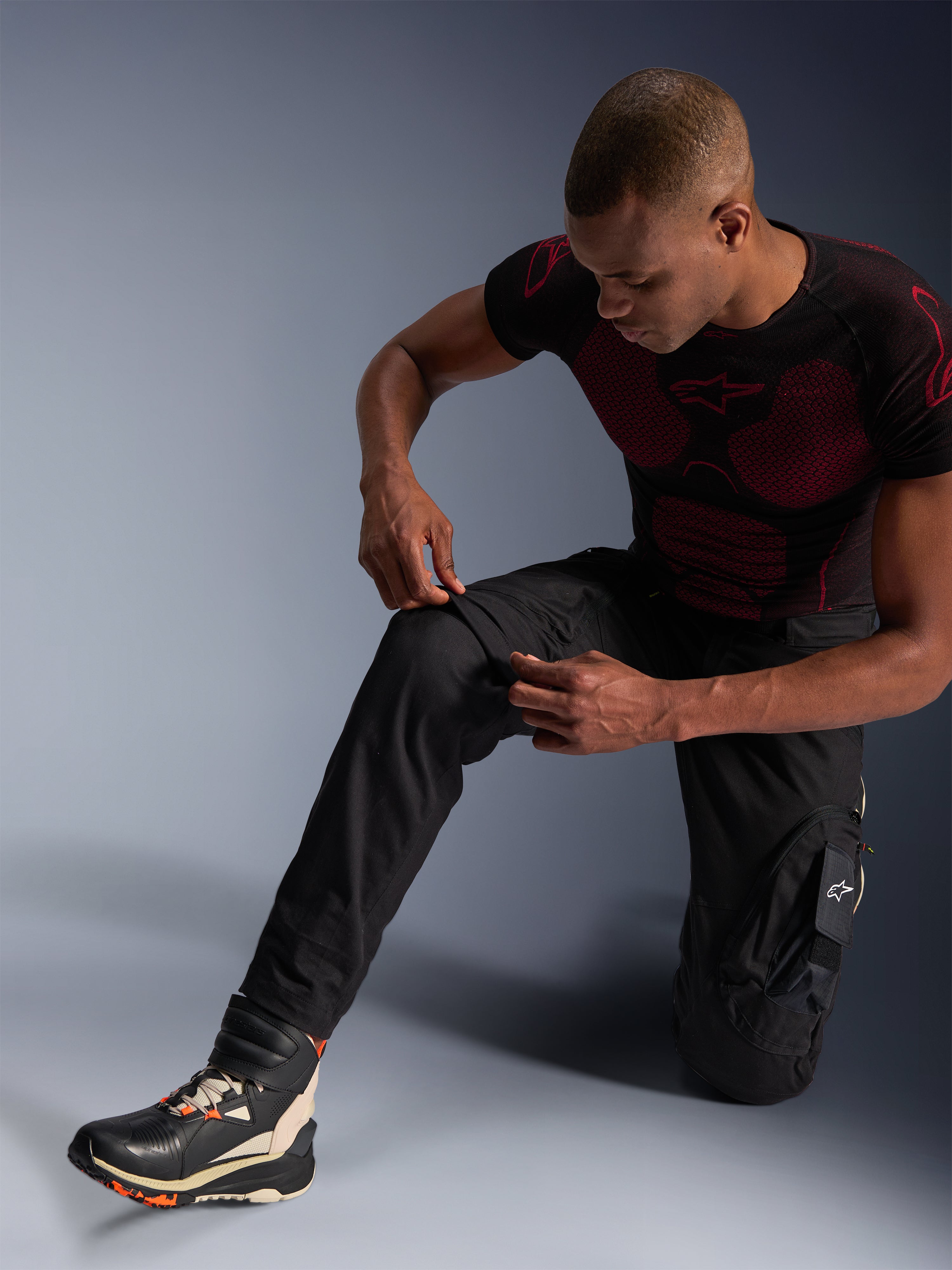 Pantalones Alpinestars Acteon, pantalones de moto urbanos negros, usados por un hombre arrodillado ajustando la zona de la rodilla, con bolsillos tipo cargo y ajuste entallado, combinados con una capa base Alpinestars negra y roja y calzado de conducción