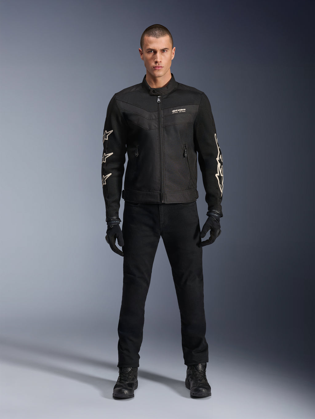 Modelo masculino con la chaqueta Alpinestars T-Dyno Air, negra, chaqueta textil de moto ligera con malla 3D transpirable, logos de estrellas blancas en las mangas, combinada con pantalones, guantes y botas negros, vista frontal de pie