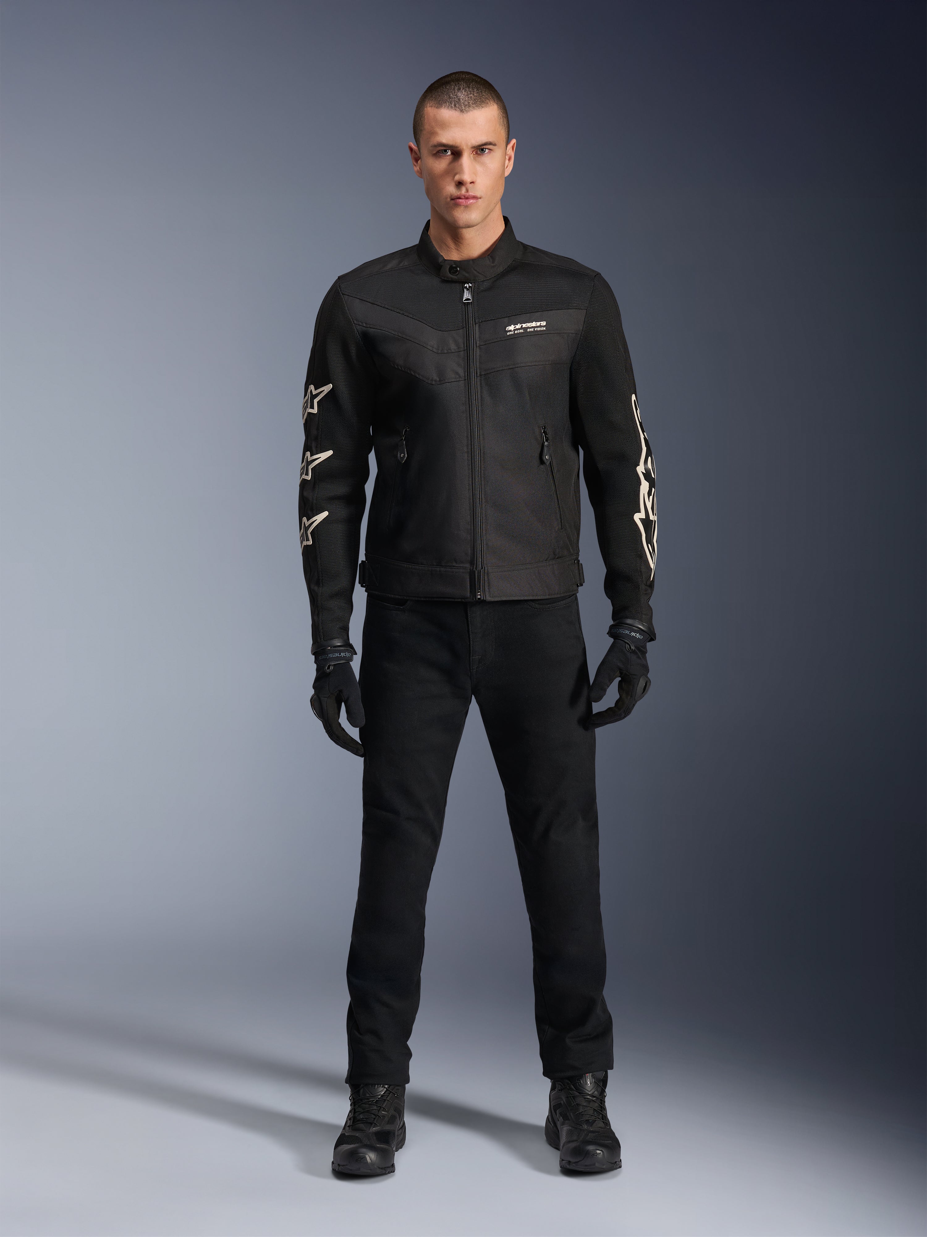 Modelo masculino con la chaqueta Alpinestars T-Dyno Air, negra, chaqueta textil de moto ligera con malla 3D transpirable, logos de estrellas blancas en las mangas, combinada con pantalones, guantes y botas negros, vista frontal de pie