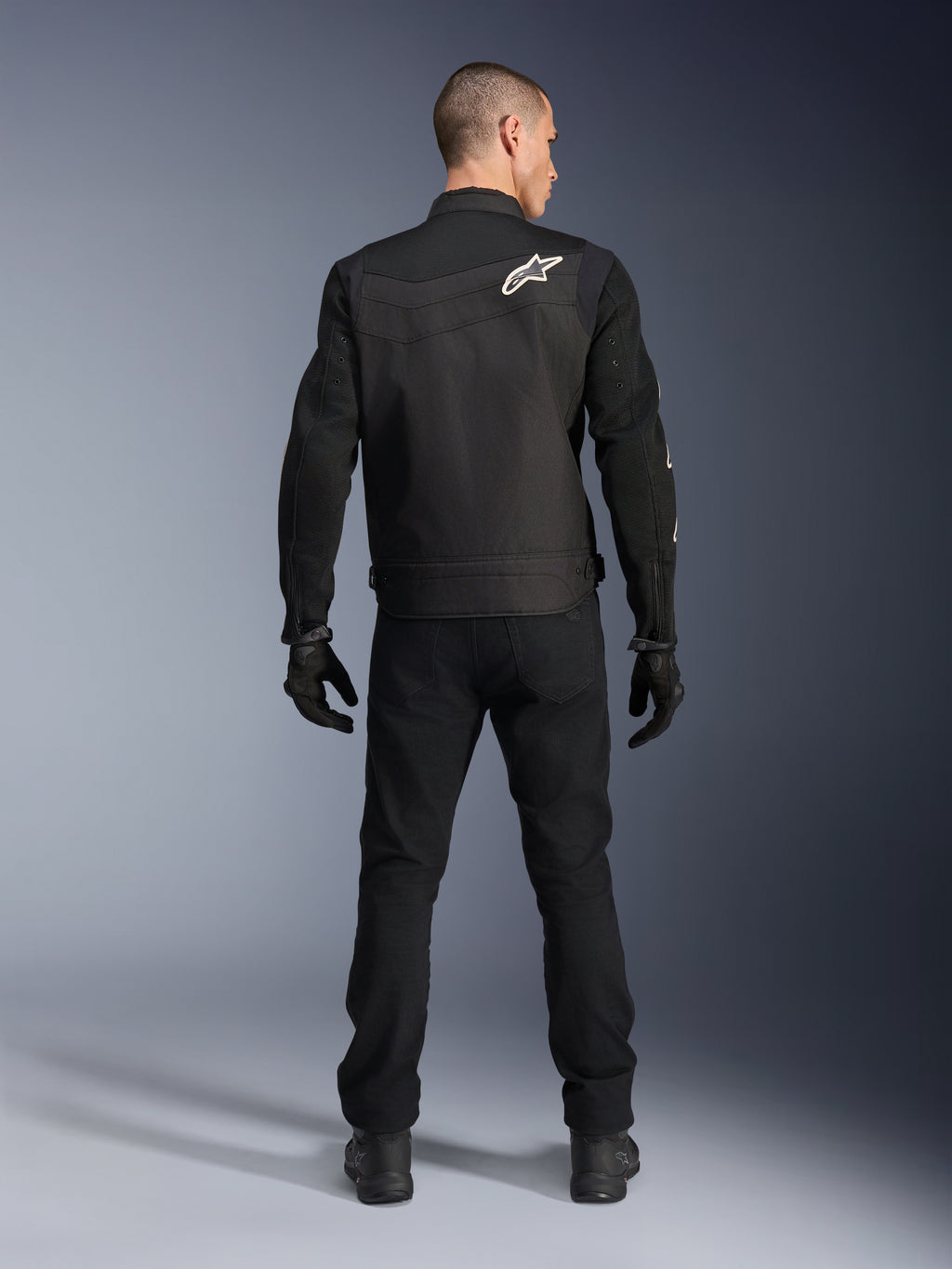 Chaqueta Alpinestars T-Dyno Air, negra, vista posterior en modelo, chaqueta textil de moto con paneles de malla 3D y un logotipo de estrella blanca en el hombro, usada con pantalones y guantes negros, conjunto de conducción urbana moderna