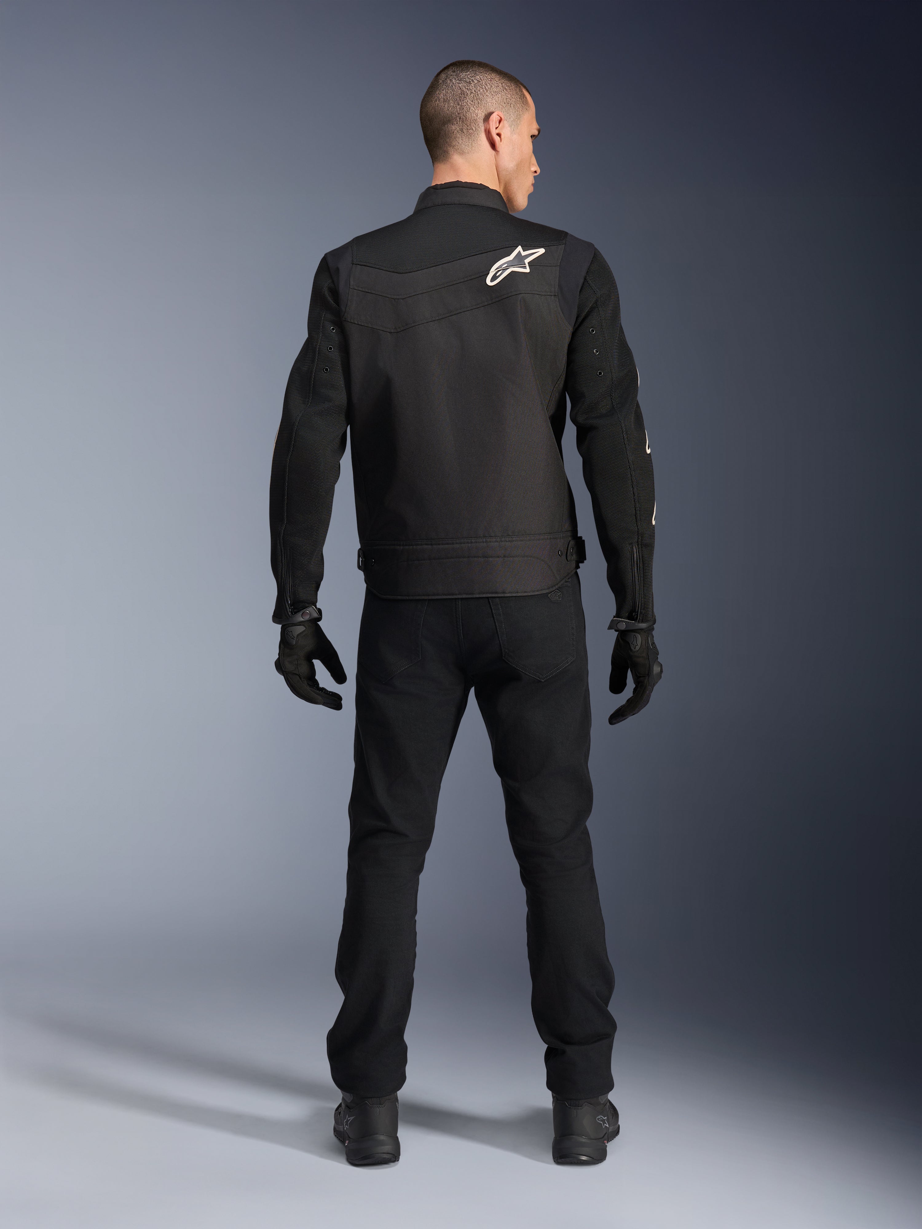 Chaqueta Alpinestars T-Dyno Air, negra, vista posterior en modelo, chaqueta textil de moto con paneles de malla 3D y un logotipo de estrella blanca en el hombro, usada con pantalones y guantes negros, conjunto de conducción urbana moderna