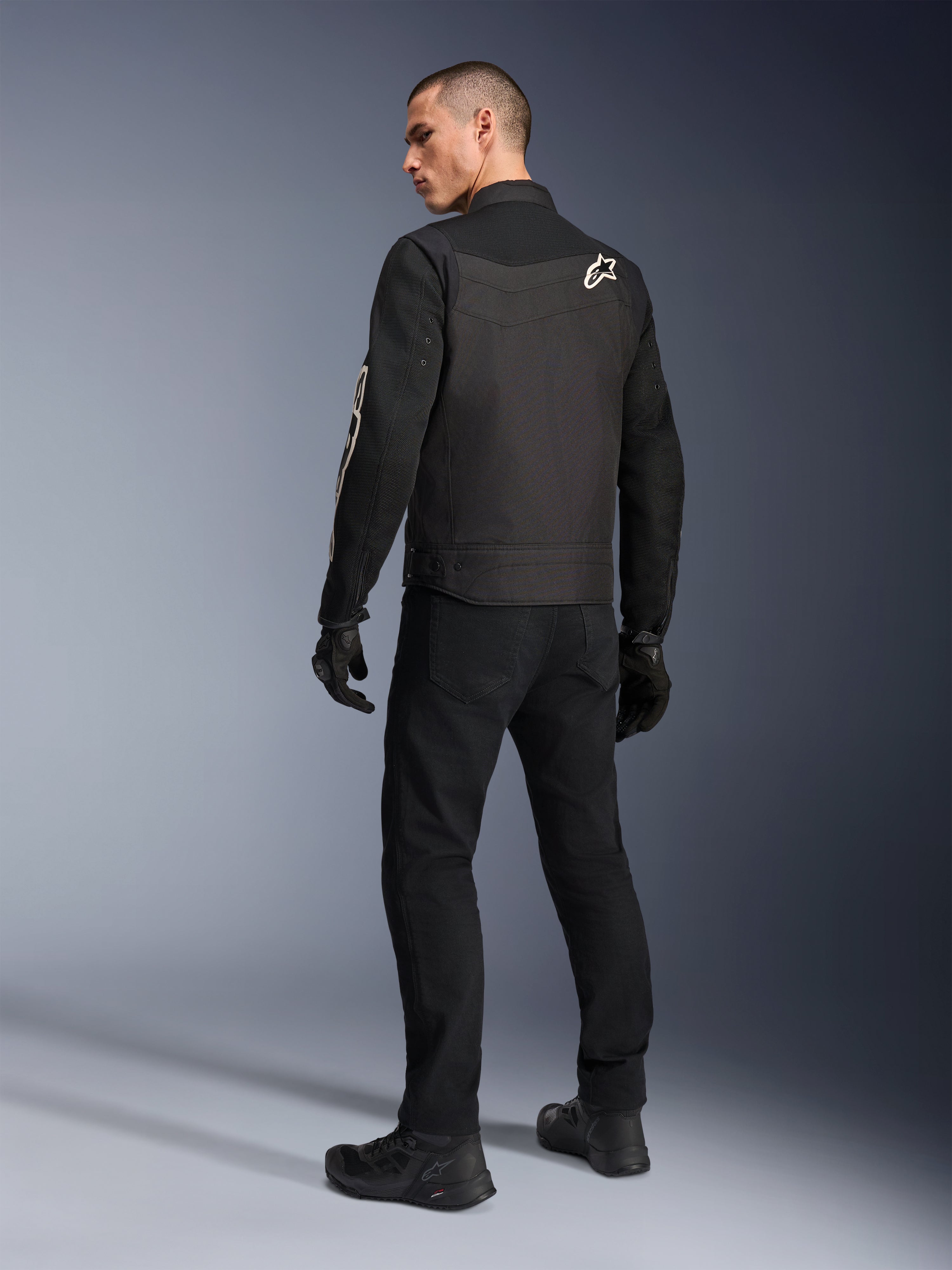 Chaqueta Alpinestars T-Dyno Air, negra, vista posterior de modelo usando una chaqueta de moto textil transpirable, con el logotipo blanco de Alpinestars en el hombro, combinada con pantalones y guantes negros, estilo de conducción urbana