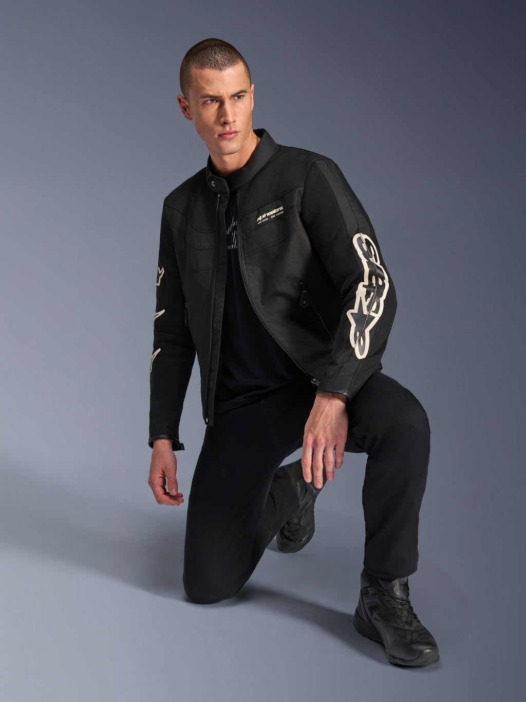 Modelo masculino vistiendo la chaqueta Alpinestars T-Dyno Air, chaqueta textil de moto negra con grandes logotipos blancos en las mangas, combinada con pantalones negros y botas de conducción, diseño moderno de inspiración vintage con paneles de malla 3D.