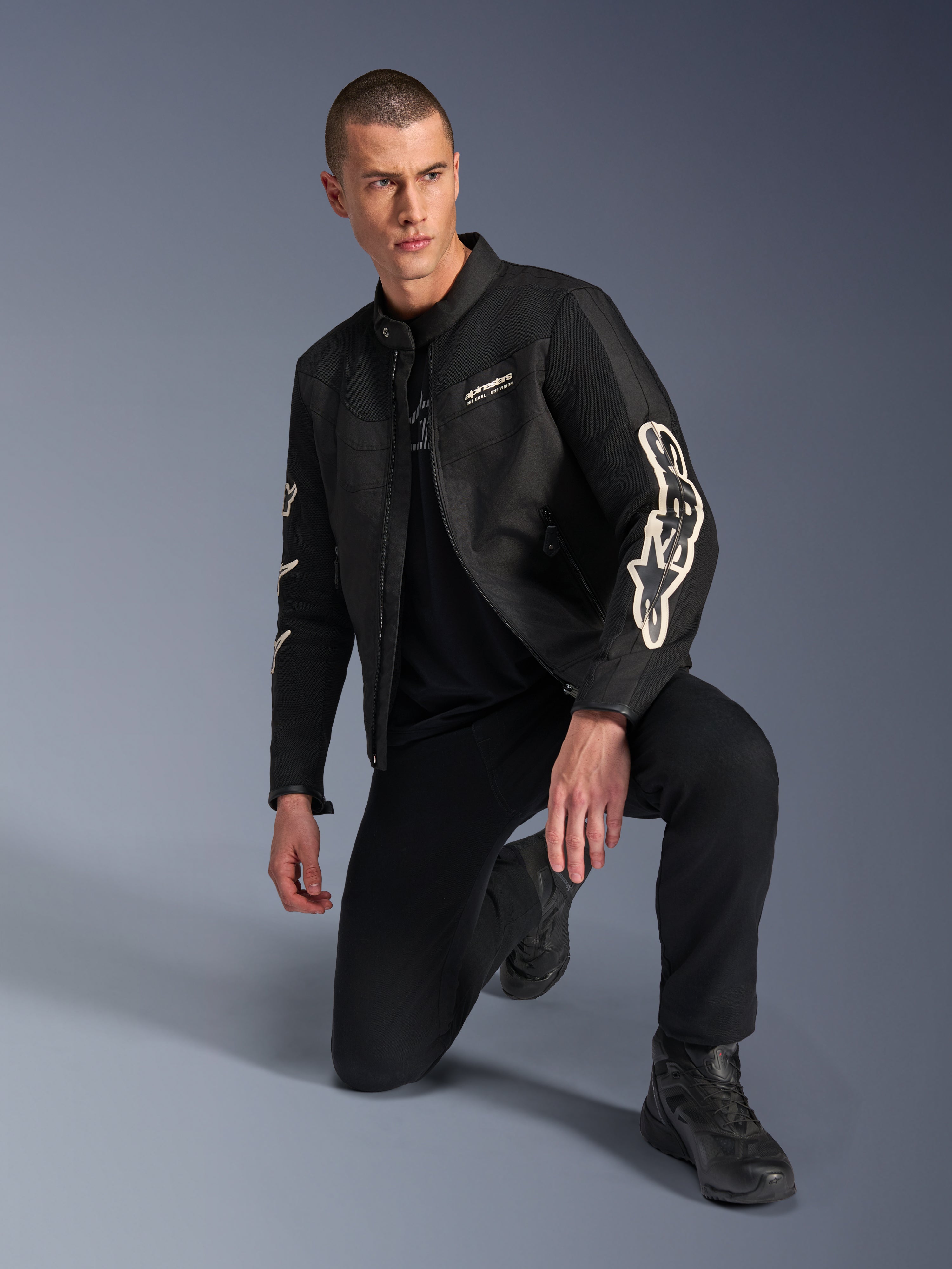 Modelo masculino vistiendo la chaqueta Alpinestars T-Dyno Air, chaqueta textil de moto negra con grandes logotipos blancos en las mangas, combinada con pantalones negros y botas de conducción, diseño moderno de inspiración vintage con paneles de malla 3D.