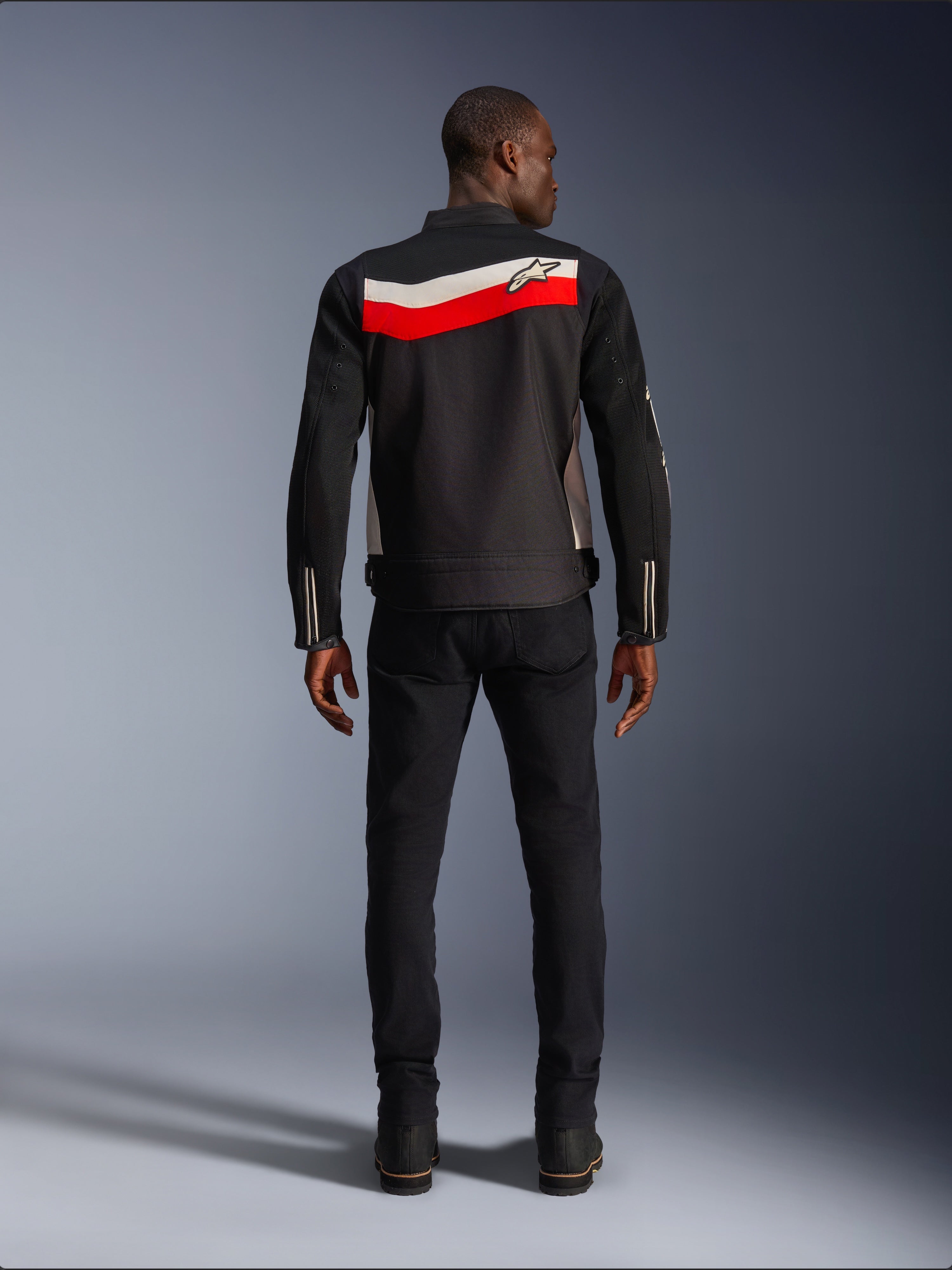 Chaqueta Alpinestars T-Dyno Air, chaqueta de moto textil para hombre, vista trasera, negra con franjas horizontales en rojo brillante y crudo en la espalda, logo de Alpinestars en el canesú, combinada con pantalones y botas negras, construcción de malla 3D transpirable