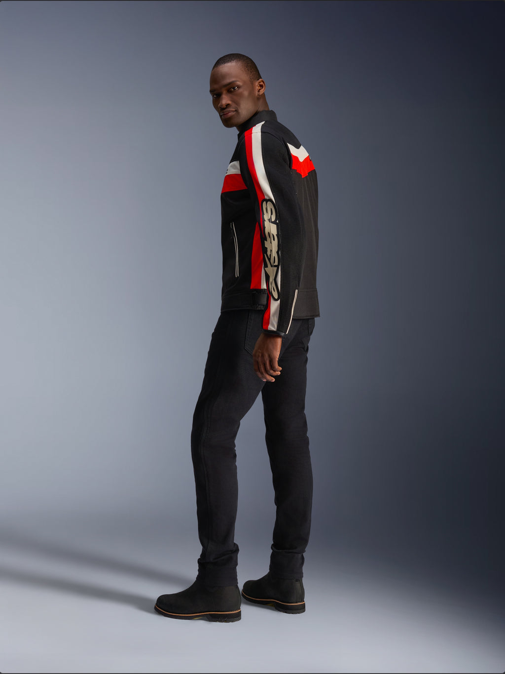 Un hombre vistiendo la chaqueta Alpinestars T-Dyno Air en negro, rojo brillante y crudo, chaqueta de moto textil de inspiración vintage con paneles de malla, marca llamativa en la manga, combinada con pantalones negros y botas para un look de motociclista moderno