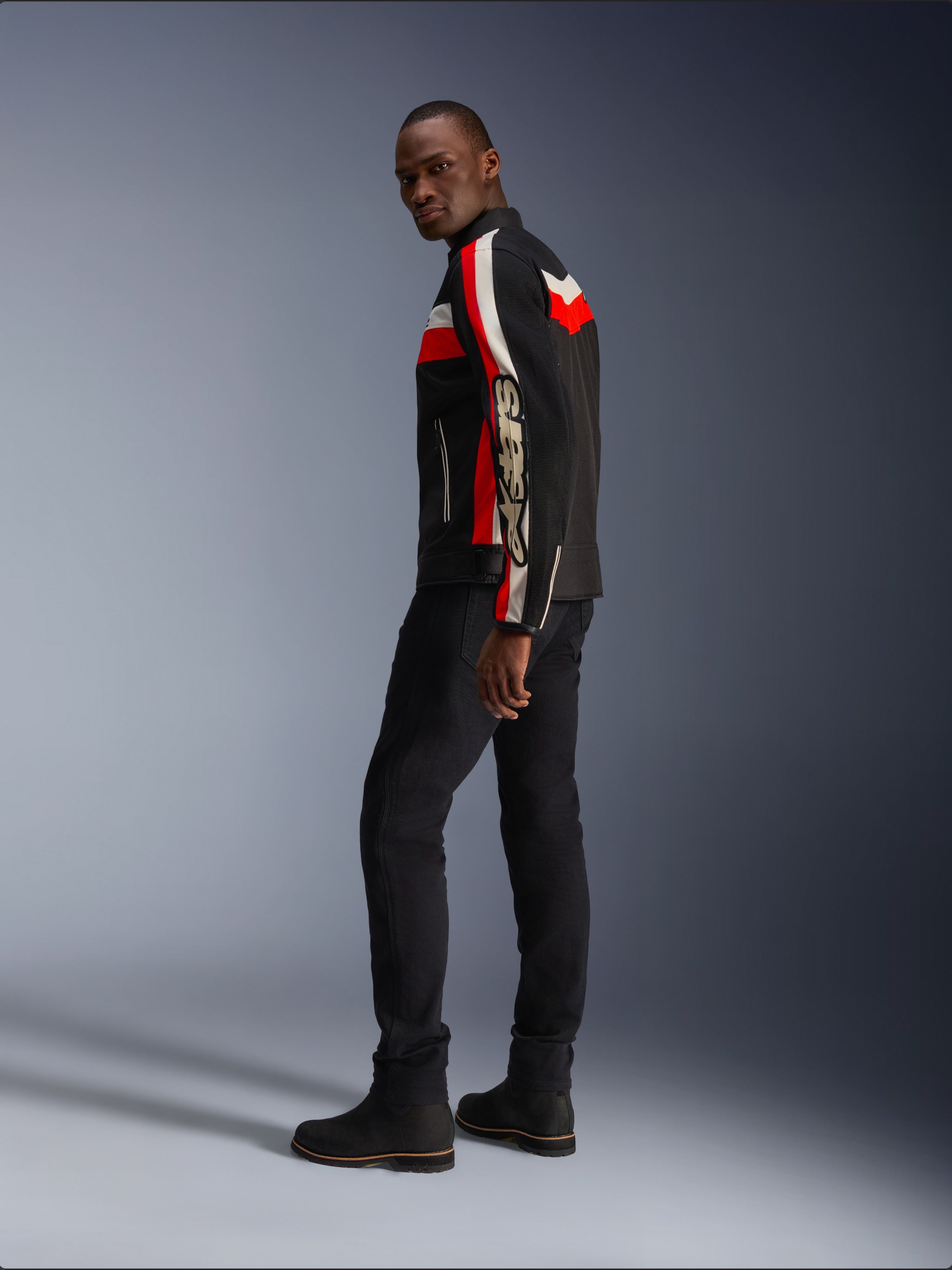 Un hombre vistiendo la chaqueta Alpinestars T-Dyno Air en negro, rojo brillante y crudo, chaqueta de moto textil de inspiración vintage con paneles de malla, marca llamativa en la manga, combinada con pantalones negros y botas para un look de motociclista moderno