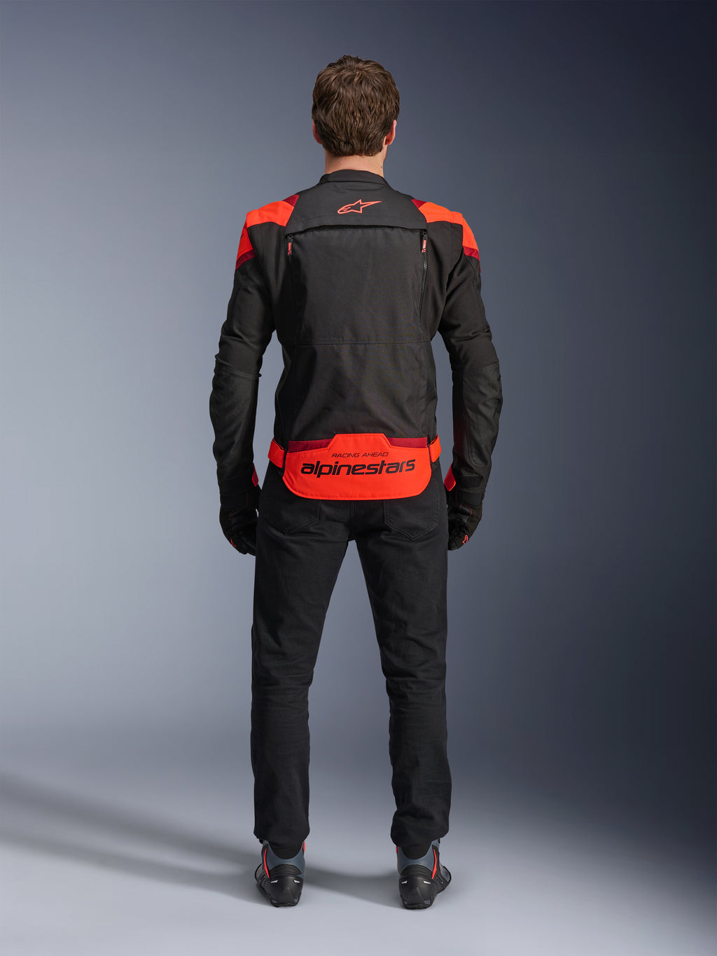 Chaqueta Alpinestars T-Stunt Air, negra con detalles en rojo flúor y rojo oscuro, vista posterior en modelo, combinada con pantalones y botas de moto negros, con un llamativo logotipo de Alpinestars en el panel inferior de la espalda, diseño modular con construcción de malla para ventilación.