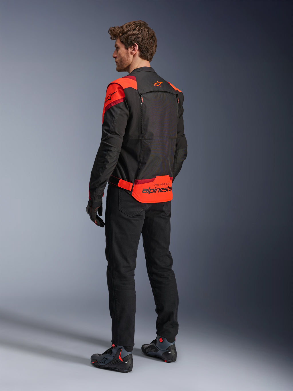 Hombre con la chaqueta Alpinestars T-Stunt Air vista desde atrás, negro, rojo fluo y rojo oscuro, chaqueta de moto textil con paneles de malla y diseño de corte deportivo, con un llamativo logotipo de Alpinestars en la parte baja de la espalda, combinada con pantalones negros y botas de motociclista