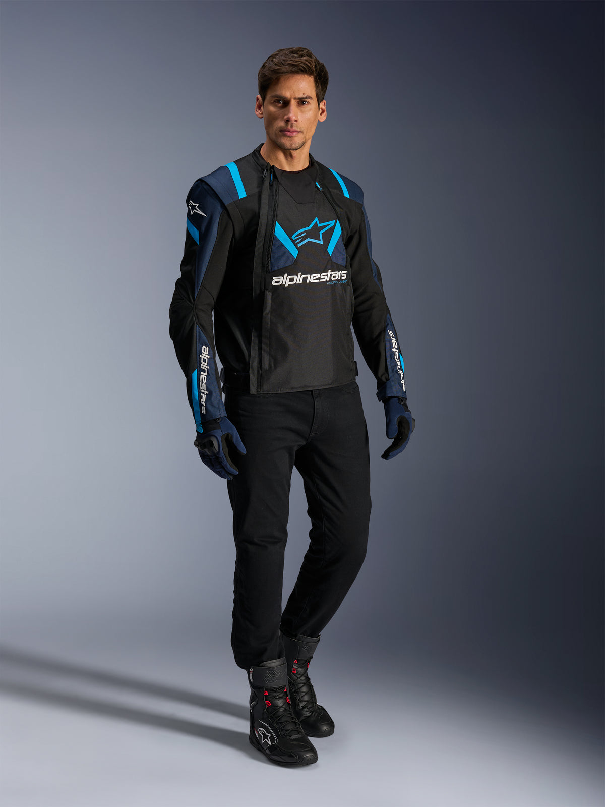 Modelo masculino con la chaqueta Alpinestars T-Stunt Air en negro, azul oscuro y azul Blithe, con paneles de malla para ventilación, combinada con pantalones negros y guantes de conducción; chaqueta de moto de corte deportivo para carretera en verano.