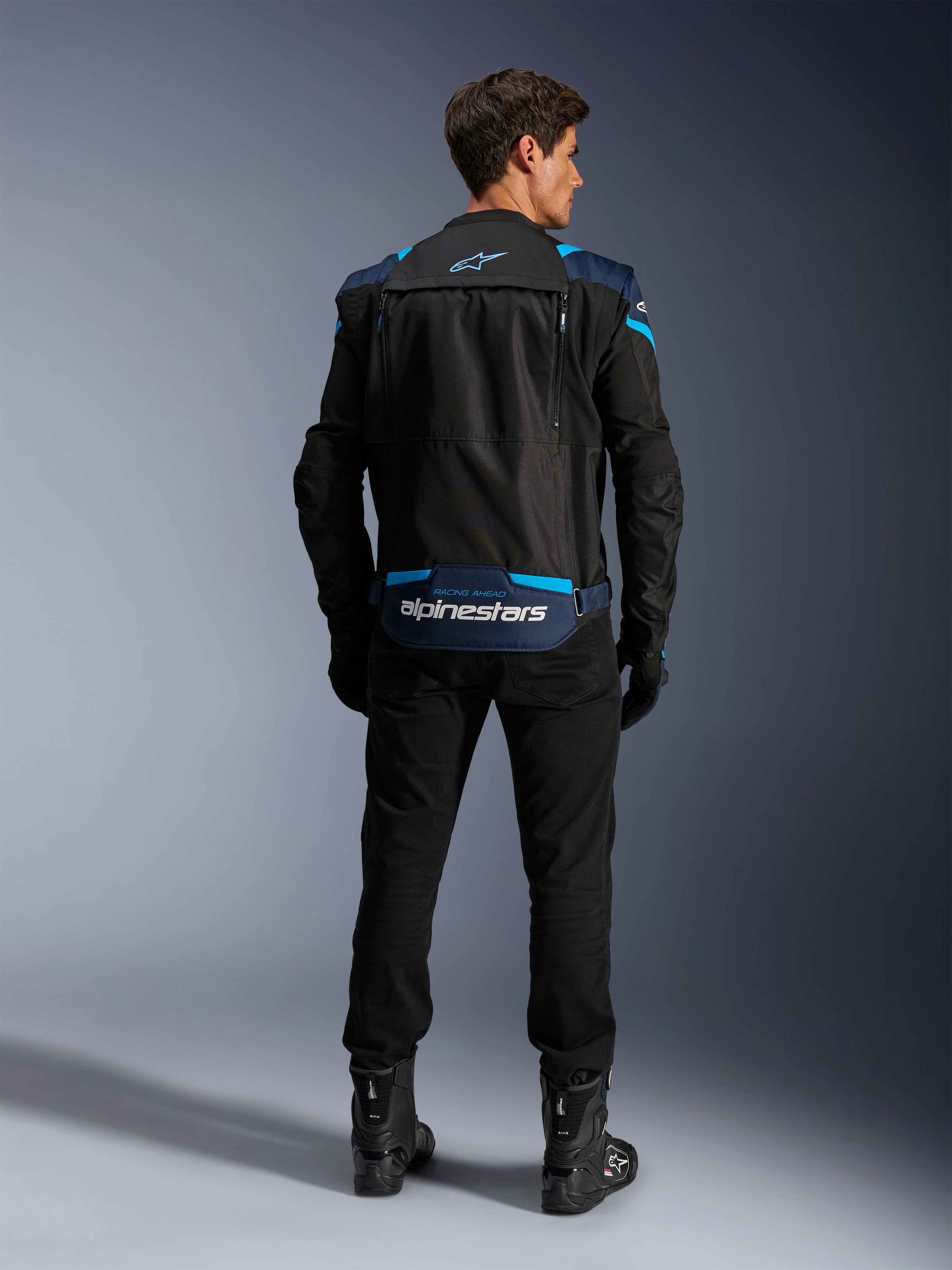 Alpinestars T-Stunt Air Jacket, chaqueta de moto textil modular, negra, azul oscuro y azul brillante, vista trasera que muestra el compartimento de almacenamiento para las mangas en la parte baja de la espalda con el logotipo blanco de Alpinestars, combinada con pantalones y botas negros