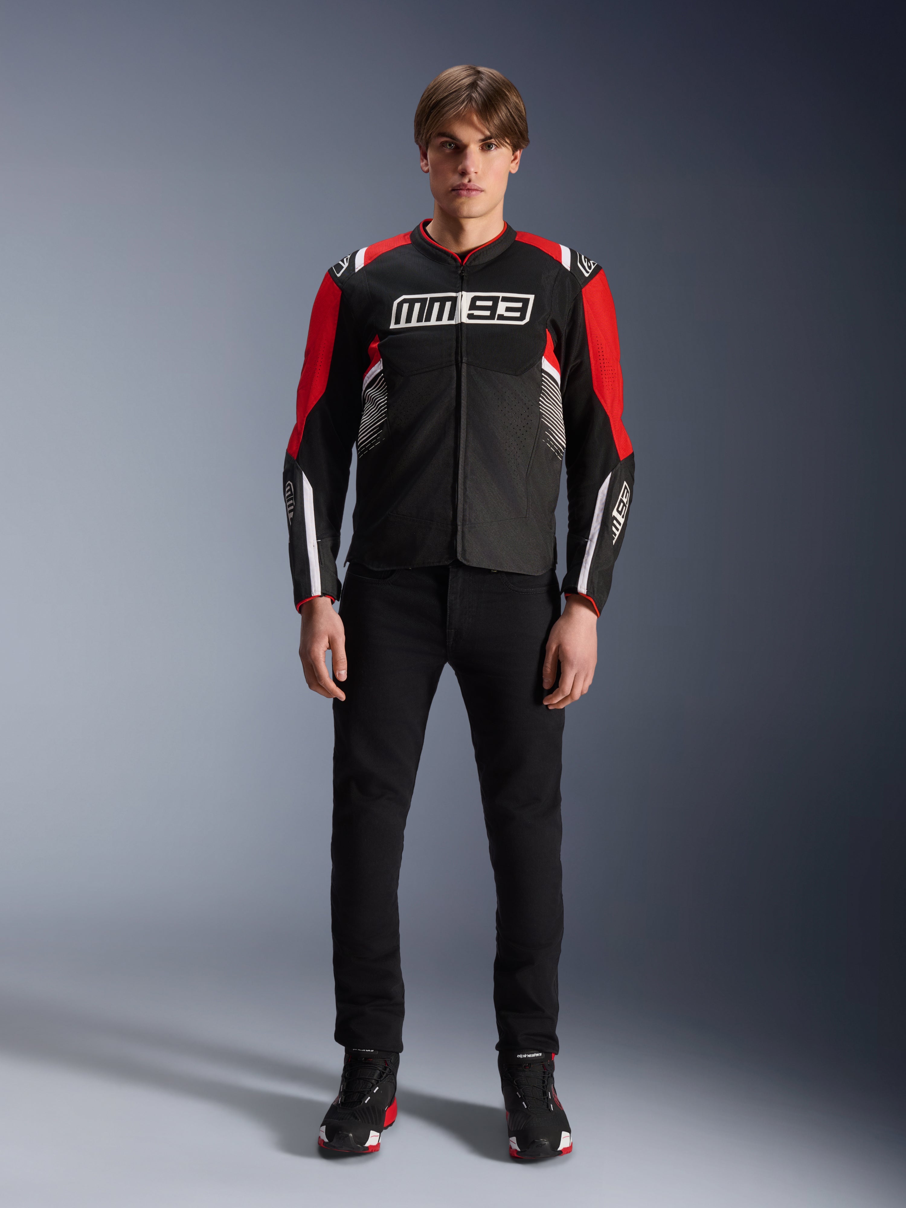 Modelo masculino vistiendo la chaqueta Alpinestars MM93 T-SPS Air V2 en negro, rojo brillante y blanco, con el logotipo MM93 de Marc Márquez en el pecho, paneles de malla para el flujo de aire, combinada con pantalones negros y zapatillas en un estudio.