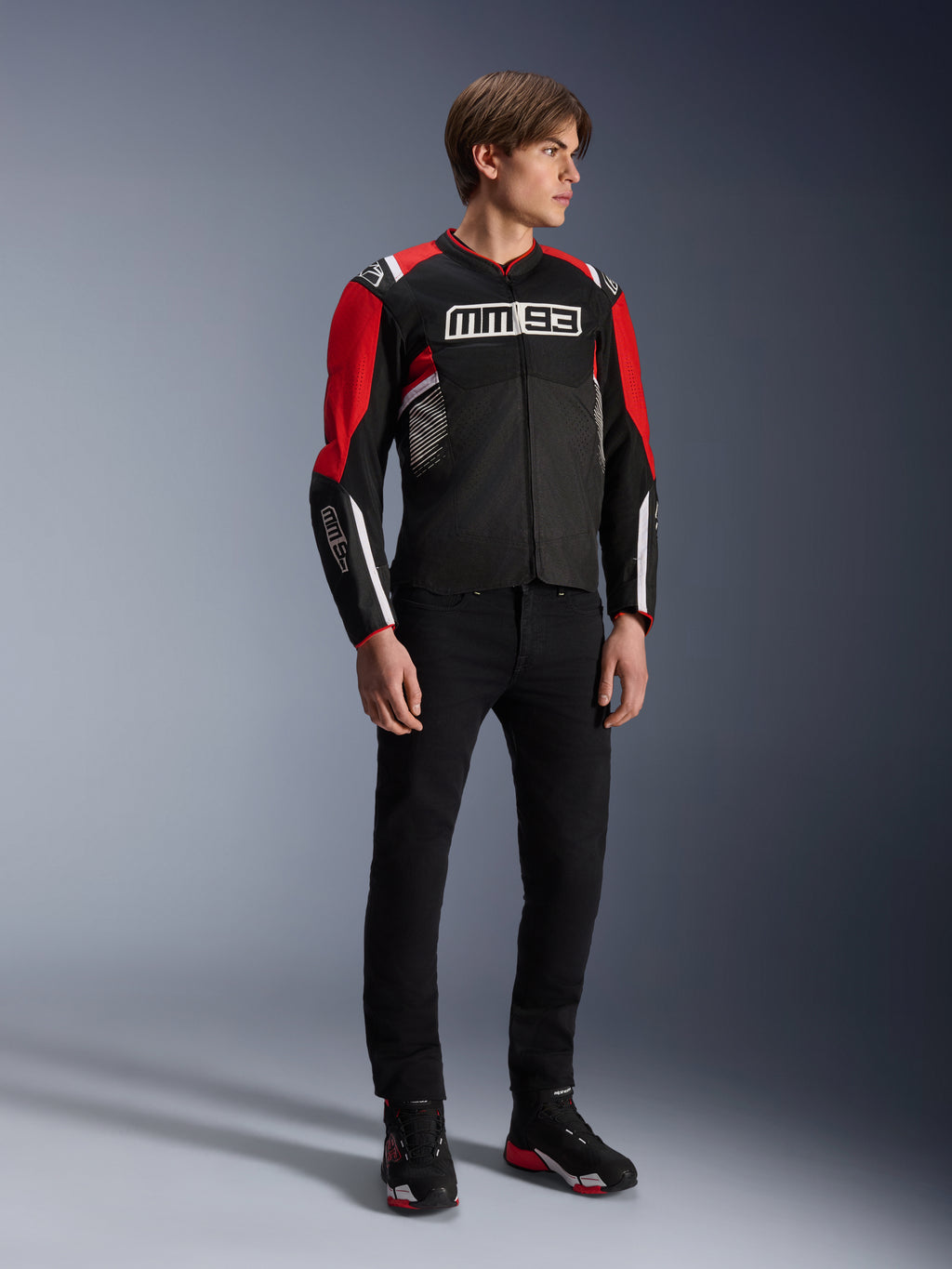 Modelo masculino vistiendo la chaqueta Alpinestars MM93 T-SPS Air v2 en negro, rojo brillante y blanco, con logotipos MM93 destacados en el pecho y las mangas, combinada con pantalones negros y botas de moto; estilo urbano deportivo con paneles de malla visibles para el flujo de aire.
