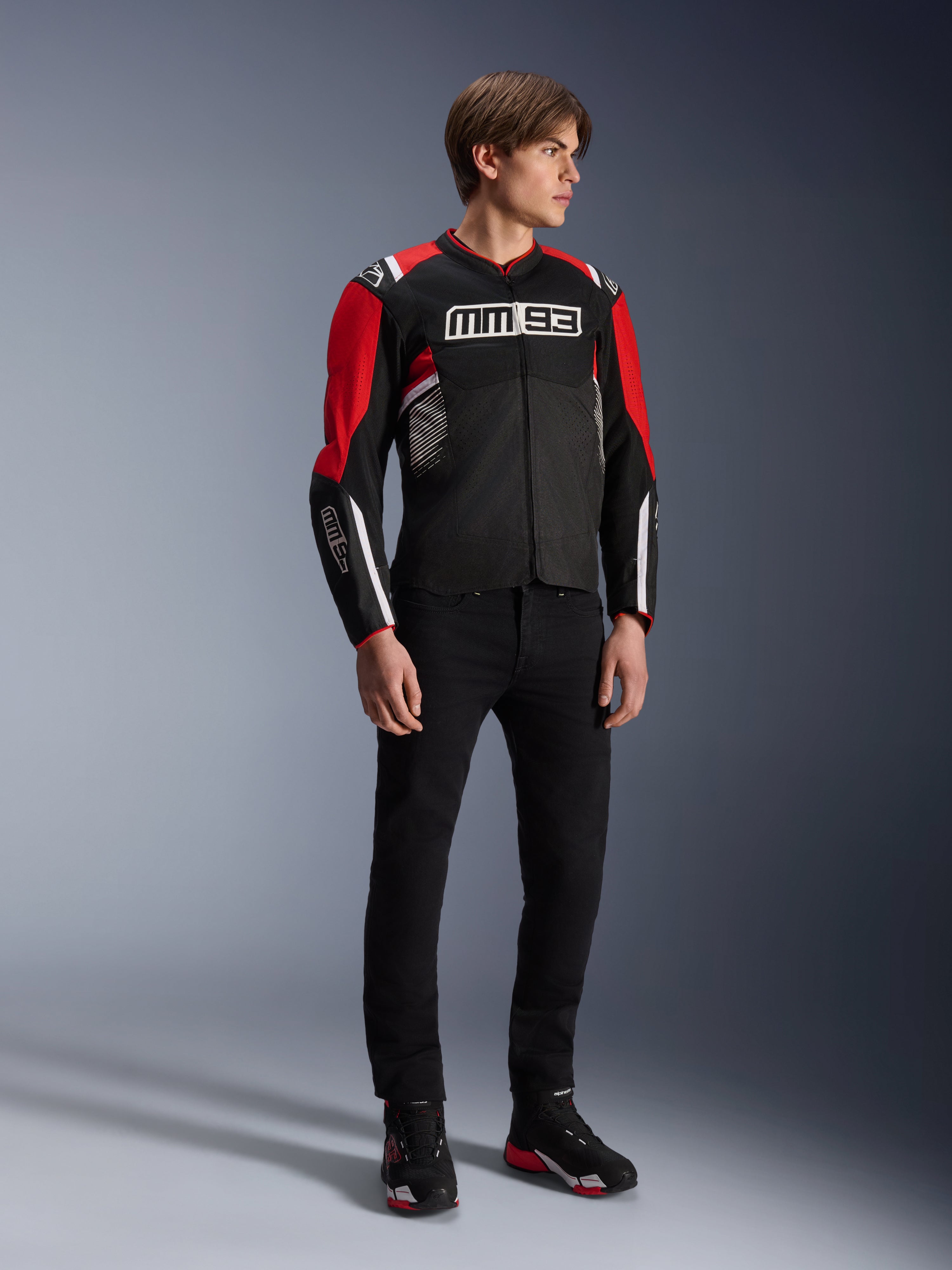 Modelo masculino vistiendo la chaqueta Alpinestars MM93 T-SPS Air v2 en negro, rojo brillante y blanco, con logotipos MM93 destacados en el pecho y las mangas, combinada con pantalones negros y botas de moto; estilo urbano deportivo con paneles de malla visibles para el flujo de aire.