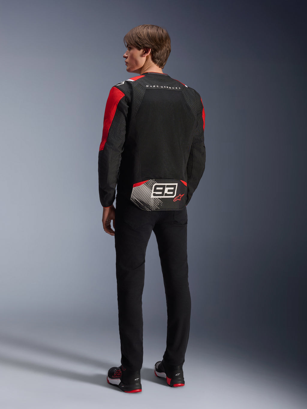 Modelo de espaldas con la chaqueta Alpinestars MM93 T-SPS Air V2, negra con detalles en rojo brillante y blanco, con el texto 'MARC MARQUEZ' y un logotipo '93' grande en la parte trasera, construcción de malla textil para ventilación, combinada con pantalones negros y calzado de moto a juego en negro y rojo