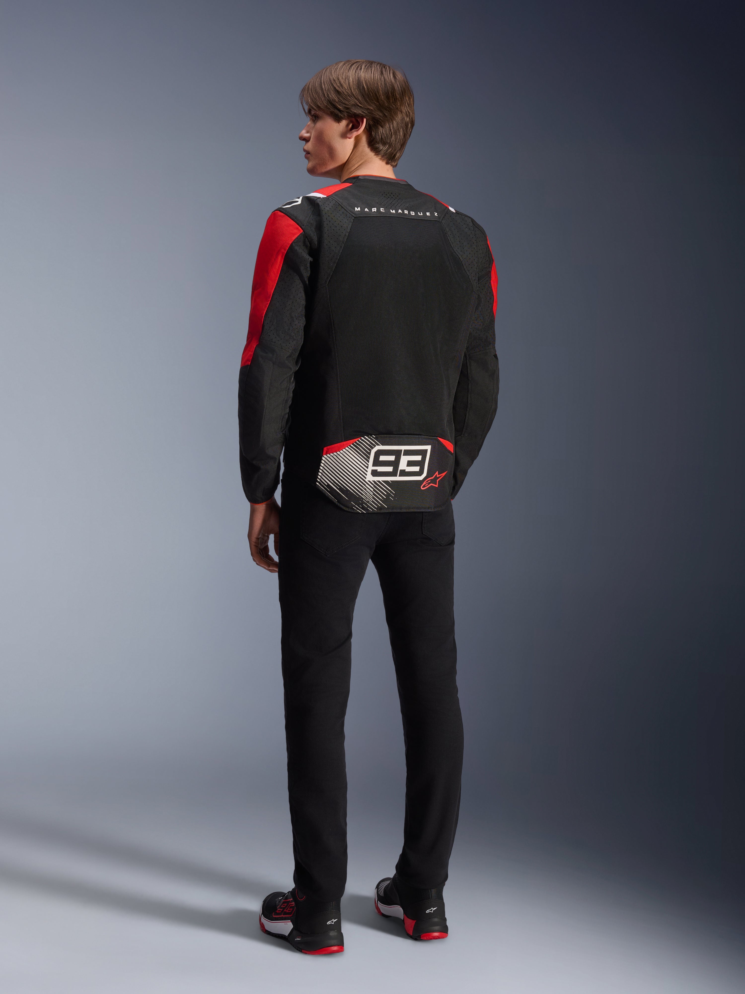 Modelo de espaldas con la chaqueta Alpinestars MM93 T-SPS Air V2, negra con detalles en rojo brillante y blanco, con el texto 'MARC MARQUEZ' y un logotipo '93' grande en la parte trasera, construcción de malla textil para ventilación, combinada con pantalones negros y calzado de moto a juego en negro y rojo