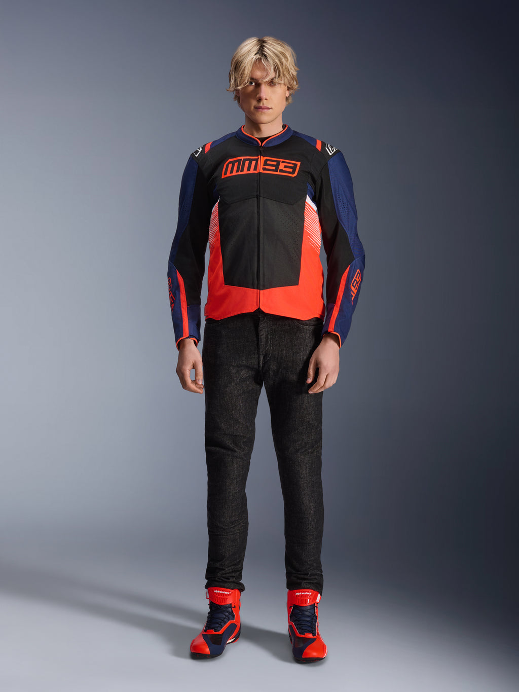 Chaqueta Alpinestars MM93 T-SPS Air V2, azul marino y negro con detalles en rojo flúor, con el icónico logotipo MM93 en el pecho, combinada con pantalones de conducción negros y botas azules y rojas a juego, atuendo de moto urbano